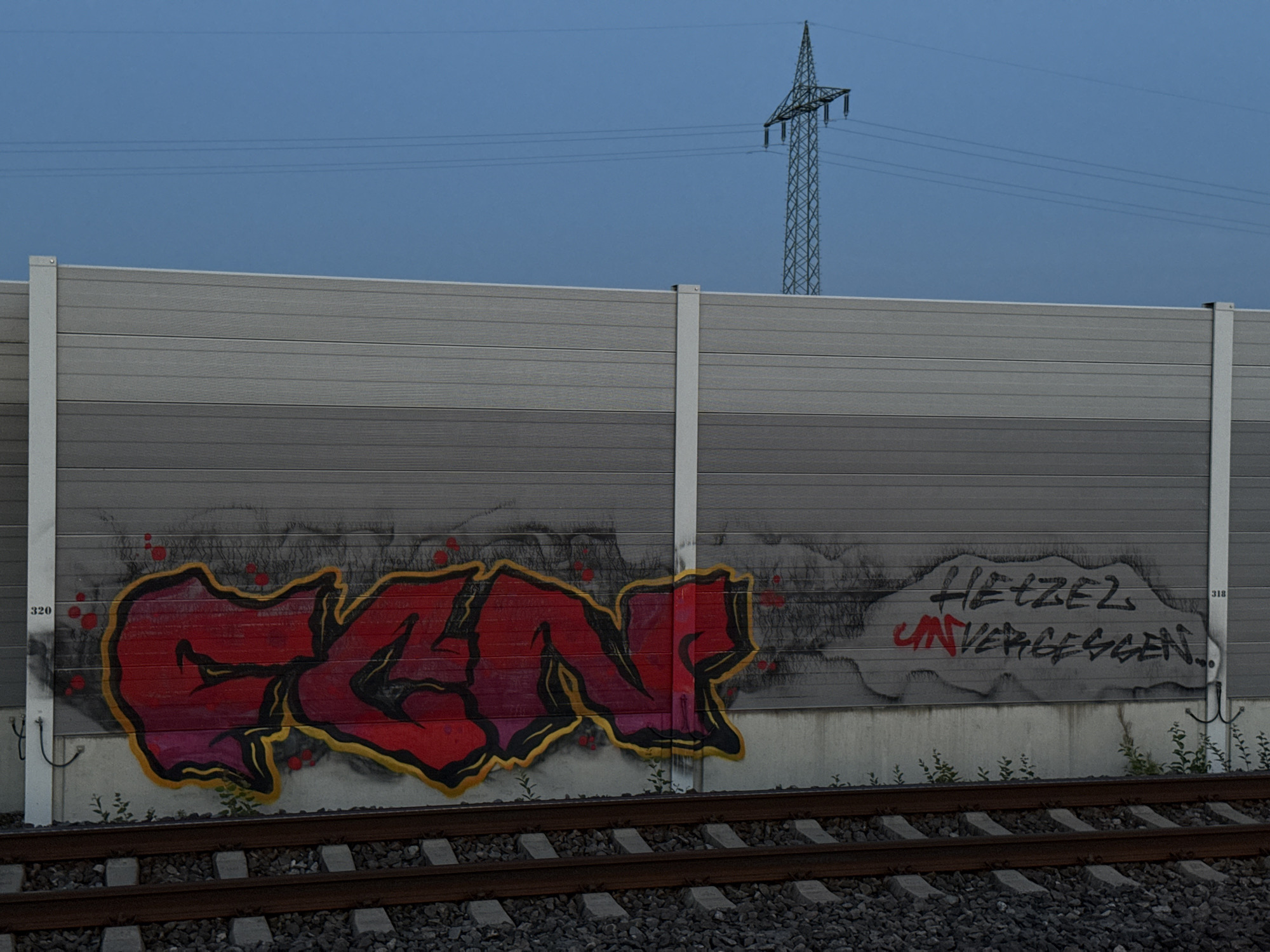 The image depicts a section of a modern, grey, horizontally slatted sound barrier erected alongside a railway track. The barrier is divided into two horizontal sections, with the upper section being a lighter shade of grey than the lower section. Graffiti adorns the lower portion of the barrier. On the left side of the barrier, large, bold red graffiti with a yellow and pink outline spells out what appears to be "COW" or similar lettering. To the right of this, within a grey cloud-shaped spray-painted area, is further graffiti in black lettering reading "HETZEL" and below it, in red, "UNVERGESSSEN." A small white marker labeled "320" is visible to the left of the red graffiti, and another labeled "318" is to the right of the cloud-shaped graffiti.
In the background, a clear, deep blue sky is visible, with a single electrical pylon and overhead power lines stretching across the horizon. The railway track in the foreground consists of weathered brown wooden sleepers and dark grey ballast. Small green plants are growing at the base of the sound barrier. The overall scene suggests an urban or industrial fringe setting, consistent with areas found in Forchheim, Germany, where such infrastructure is common. There are no people or animals present in the image, and the focus is on the static elements of the environment and the graffiti.