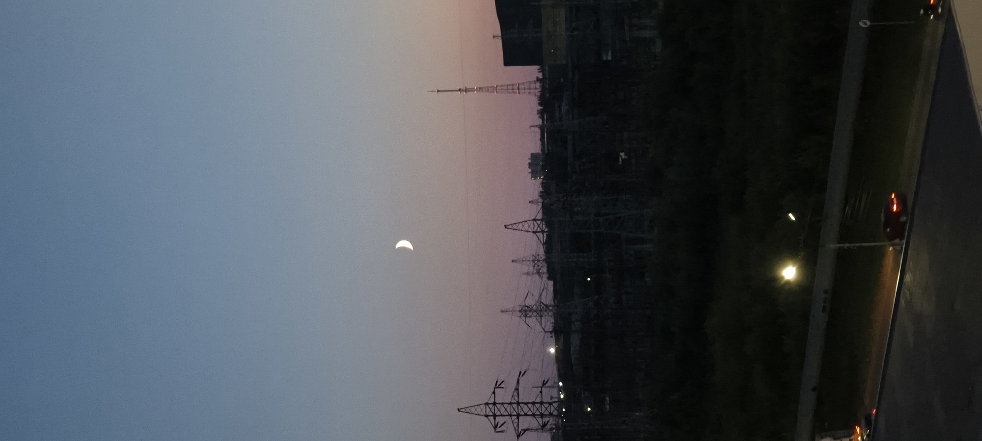 A crescent moon hangs in a gradient sky of deep blue and pale pink over a landscape of power lines and towers. The power infrastructure dominates the mid-ground, with tall, lattice-like towers and numerous wires stretching across the scene. In the far distance, the silhouette of buildings and a tall antenna structure are visible. The foreground shows a dark, tree-covered area, and a road with a single streetlamp illuminating a red car parked on the right side of the frame. The overall impression is of an industrial or semi-industrial area at dusk or dawn.