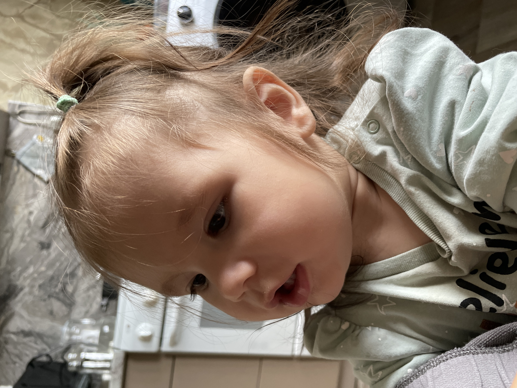 A close-up portrait captures a young child, likely a toddler, with their head positioned in the upper left of the frame, angled downwards. The child has fair skin and light brown hair, pulled into a small, somewhat dishevelled ponytail on top of their head, secured with a green hair tie. Their dark eyes are focused downwards and to the left, indicating observation. The child's mouth is slightly open, revealing some lower teeth and a hint of moisture on the lips, suggesting curiosity or speech.
The child wears a light mint green long-sleeved top or onesie, adorned with faint patterns that resemble stars and other abstract shapes. Partially visible in black lettering on the front of the shirt is the word segment "elieve," likely part of "believe." A layer of grey, possibly ribbed, clothing is visible beneath the mint green top around the neck and shoulder area.
The background is softly blurred, revealing an indoor, domestic setting, possibly a kitchen or living area. Vague shapes of a white appliance with a black circular component can be discerned in the upper right. The scene is set indoors, likely in a home in Izhevsk, Russia. The overall impression is one of candid youthful inquisitiveness.