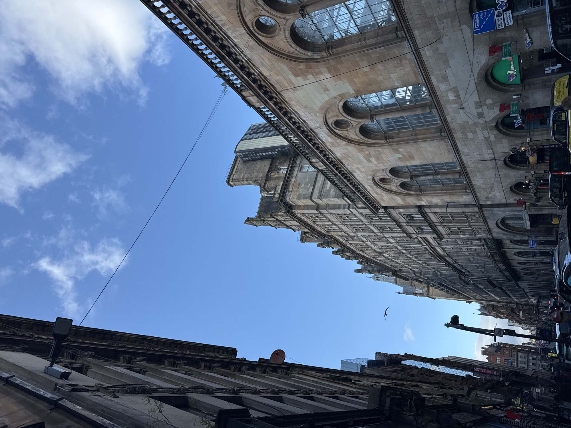 The image shows a view looking up at buildings in the city of Glasgow, Scotland, United Kingdom. The architecture appears to be historic, with ornate stonework and large arched windows on the building to the right. The building on the left has a more modern facade with vertical windows. The sky above is a clear blue with a few scattered white clouds. A bird is visible in flight against the sky. Several streetlights are mounted on poles, and there are power lines crisscrossing between buildings. Traffic can be seen on the street below, including taxis. Signage for businesses such as "Reardons" and "Artio Hotel" are visible on the ground floor of some buildings. The scene depicts a typical urban environment with a mix of old and new architecture.