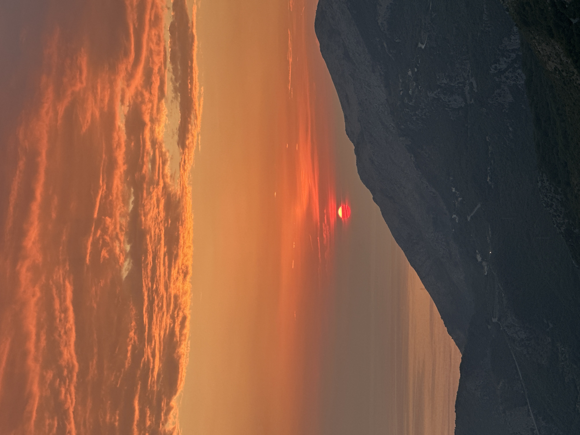 A sunset scene is captured from a vantage point above a mountainous coastline. The sun, a vibrant red orb, is partially obscured by clouds and is setting over the Adriatic Sea. The sky is a dramatic display of orange and red hues, with streaks of darker clouds adding depth and texture. In the foreground, a steep, dark mountain slope dominates the lower right of the image, sparsely dotted with lights from what appear to be scattered settlements. A winding road is visible traversing the mountainside. The sea stretches out to the left, reflecting the warm colors of the sunset. The location is specified as Đurovići, Montenegro.