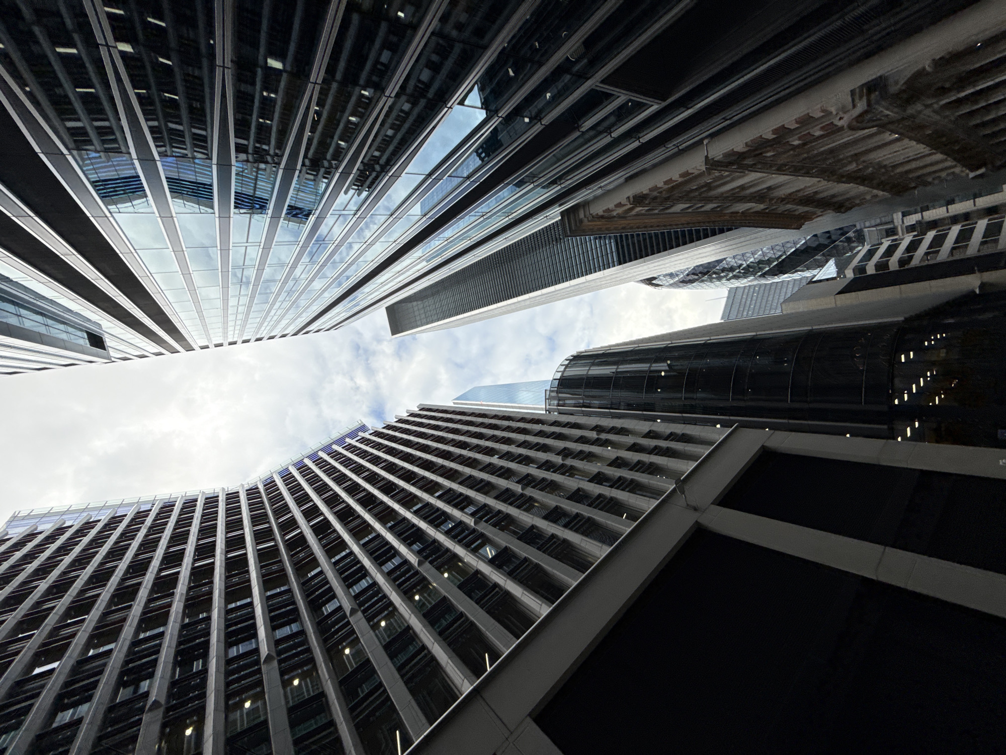 The image captures a low-angle perspective of skyscrapers in the City of London, United Kingdom. The buildings are characterized by their modern architecture, featuring extensive glass facades that reflect the cloudy sky and surrounding structures.
On the left, a large, dark building with a ribbed facade curves upwards, its windows appearing as dark rectangular openings. Adjacent to it and slightly to the right is another tall, modern building with a uniform pattern of horizontal bands of windows and darker panels.
In the center of the frame, a slender, glass-clad skyscraper with a distinctive curvilinear form rises towards the sky, appearing somewhat translucent against the overcast sky. To its right, a tall, rectangular skyscraper with a facade of alternating dark and reflective panels dominates the view. Further to the right, another building with a more traditional architectural style, featuring ornate stonework and arched windows, is visible.
The sky is overcast, with visible clouds, suggesting a daytime scene. The overall impression is one of urban density and architectural modernity, with a focus on the verticality of the cityscape. No people or vehicles are visible in the image.