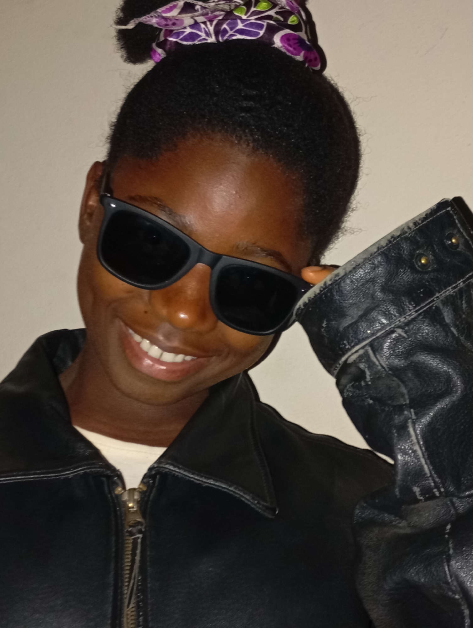 A young woman with dark skin is wearing black sunglasses and a black leather jacket. She is smiling slightly and has her hand near her sunglasses. Her hair is pulled back with a patterned purple and white headband. The background appears to be a plain, light-colored wall. There is no visual information in the image that suggests a location in Jos, Nigeria.