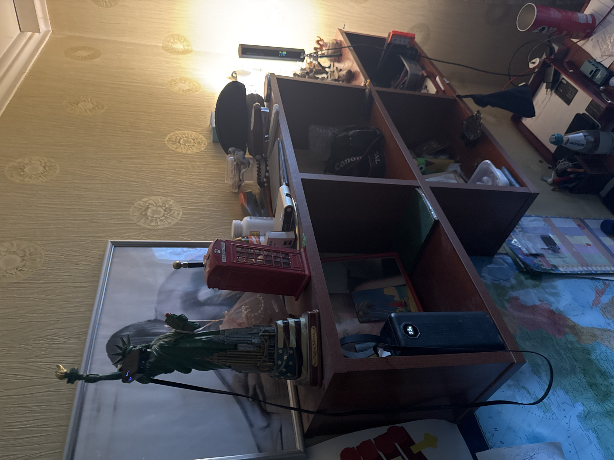 The image shows a cluttered shelf unit in what appears to be a room in Sertolovo, Russia. The shelf unit is made of dark wood and has multiple compartments. On the top shelf, there is a statue of the Statue of Liberty, a red telephone booth replica, and various bottles and containers. The middle shelf holds a black camera bag, a small tripod with figurines, and some miscellaneous items in bins. The bottom shelf is mostly obscured, but appears to contain more storage containers. Below the shelf unit, a map of the world is visible on a desk, along with a bottle of water, a small toy car, and a red Pringles can. The lighting in the room is dim, with a strong light source coming from the right, creating shadows and highlighting the dust and texture of the wallpaper.