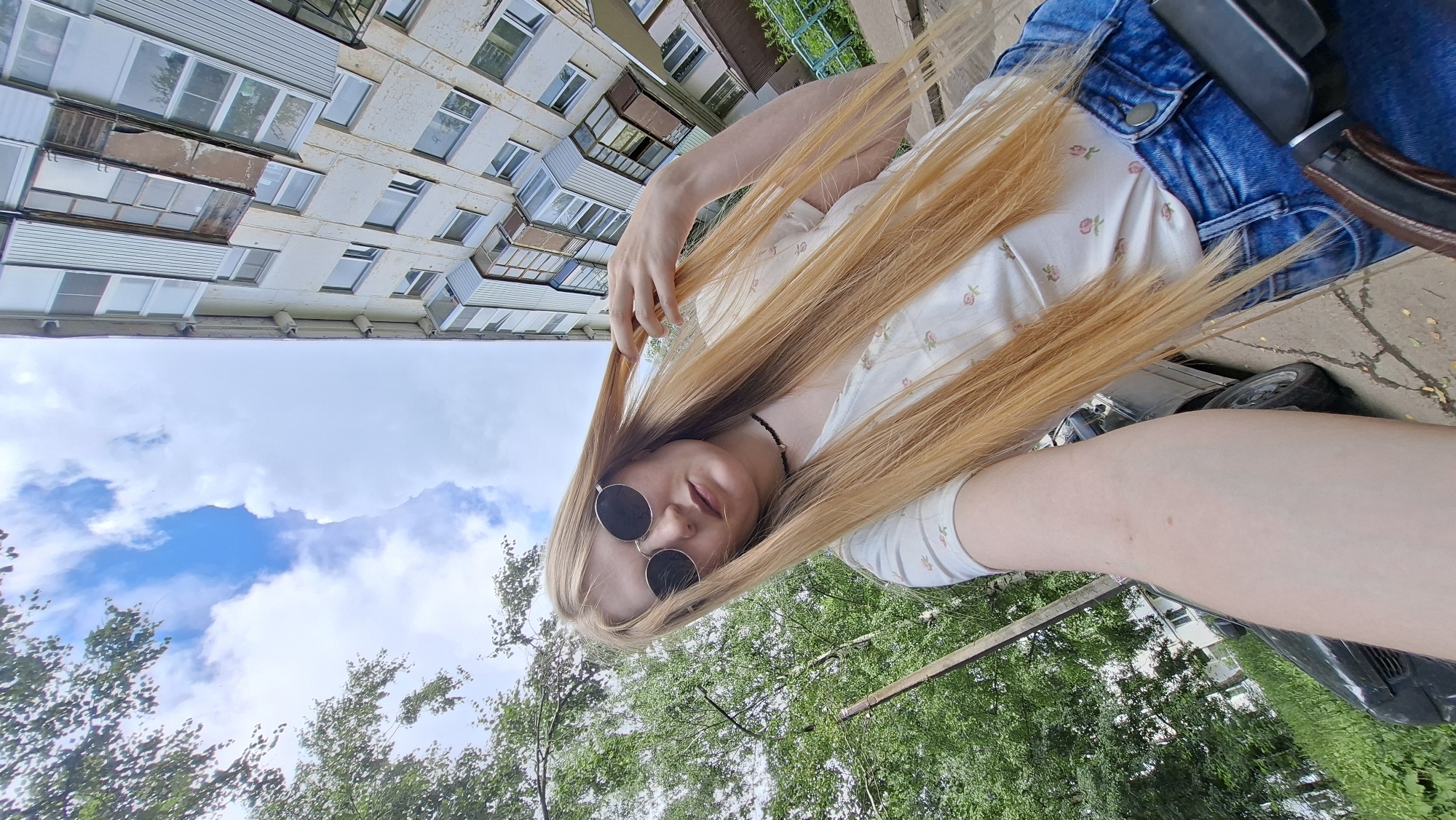 A young woman with long blonde hair and round sunglasses is taking a selfie outdoors. She is wearing a white t-shirt with a floral pattern and blue denim shorts. Her hair is partially covering her face as she touches it with her right hand. In the background, there is a tall, multi-story apartment building with balconies and windows, trees, a car parked on the street, and a cloudy sky. The image appears to be taken in the city of Chaikovsky, Russia, as indicated by the style of the building and the general urban landscape.