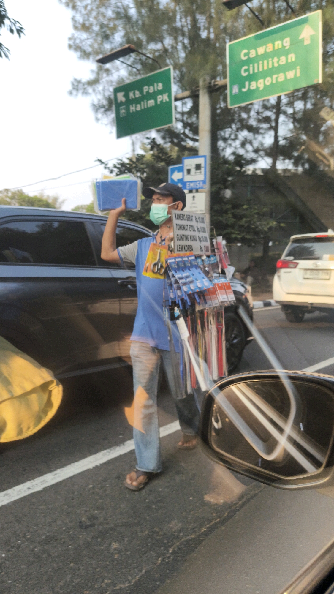 A street vendor, wearing a light blue t-shirt, blue jeans, a black cap, and a light blue face mask, stands on the asphalt road in East Jakarta, Indonesia. He is holding a stack of blue folders in his left hand and a rack of goods, including scissors, lighters, and pens, in his right hand. The vendor is positioned between two cars, a dark-colored SUV on his left and a white SUV on his right, suggesting he is operating in traffic. Traffic signs indicating directions to "Kb. Pala Halim PK" and "Cawang Cilitan Jagorawi" are visible in the background, along with trees and a cloudy sky. The scene is likely a busy intersection or highway where the vendor is soliciting sales from passing motorists.