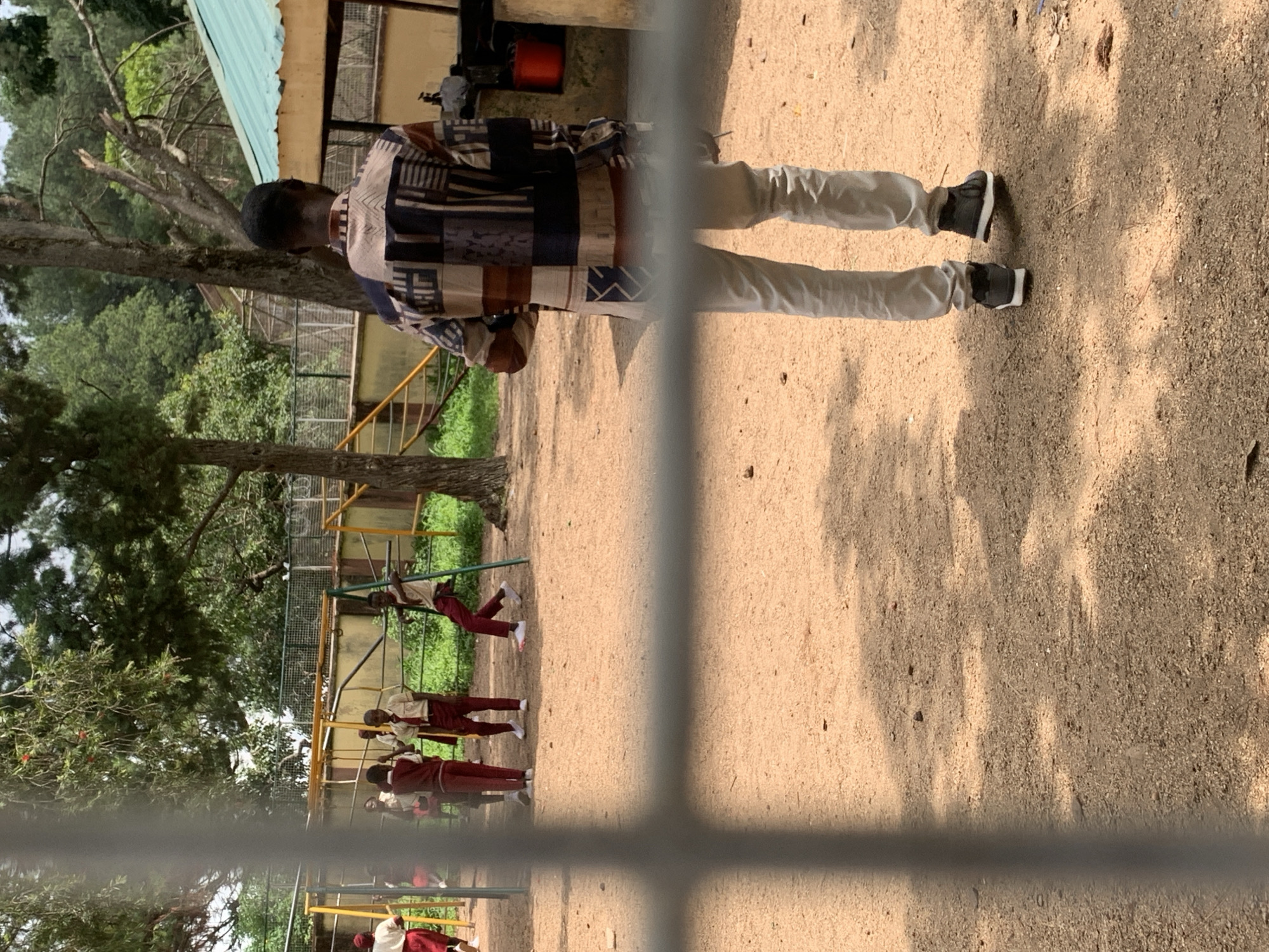 The image depicts an outdoor scene, likely a schoolyard or park, with a group of children in uniform standing near playground equipment. The children are wearing maroon and white uniforms, suggesting a school setting. One child is on a swing set. In the foreground, an adult male, viewed from behind, is wearing a patterned shirt and light-colored pants. The ground is covered with dirt and sand. The background includes trees, a building with a green roof, and a fence. The presence of the playground equipment and the children in uniform strongly suggests this is a school environment. The query about the city of Jos, Nigeria is not supported by any visual cues within the image.