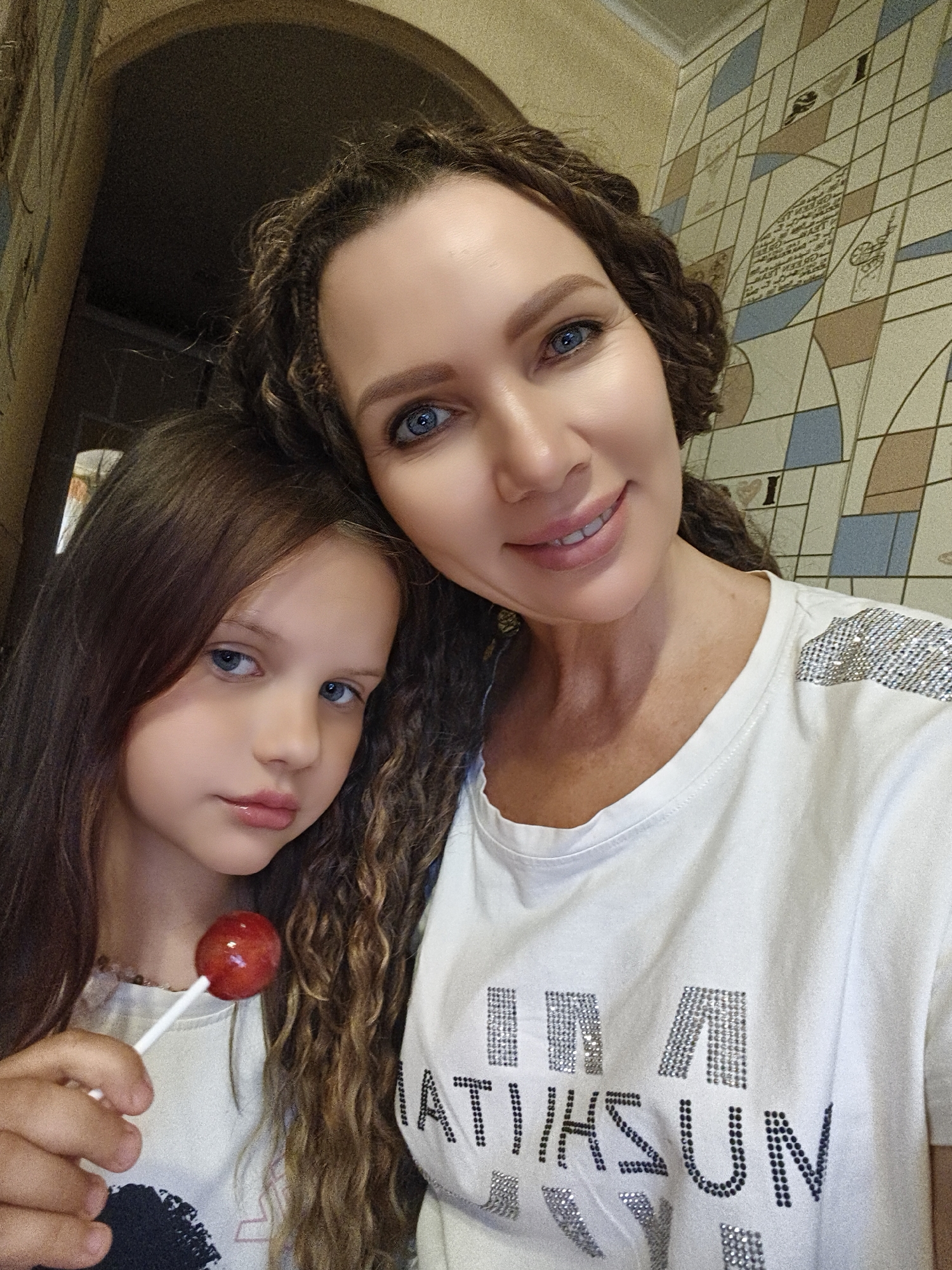 A medium shot captures two females in a close-up selfie. The female in the foreground, on the right, is an adult with long, curly brown hair and striking blue eyes. She is wearing a white t-shirt adorned with sparkling embellishments on the shoulders and across the chest, forming the words "I'M A SUN." Her mouth is slightly open in a smile, revealing her teeth, and her head is tilted slightly to the left. To her left and slightly behind is a young girl with long, straight brown hair and blue eyes. She is holding a red lollipop in her right hand, which is positioned near her mouth. The girl's expression is more subdued, with her gaze directed forward. The background appears to be an interior space with a tiled wall featuring a geometric pattern of beige, brown, and blue squares, interspersed with text and circular motifs. Part of an arched doorway or structure is visible in the upper left corner of the image.