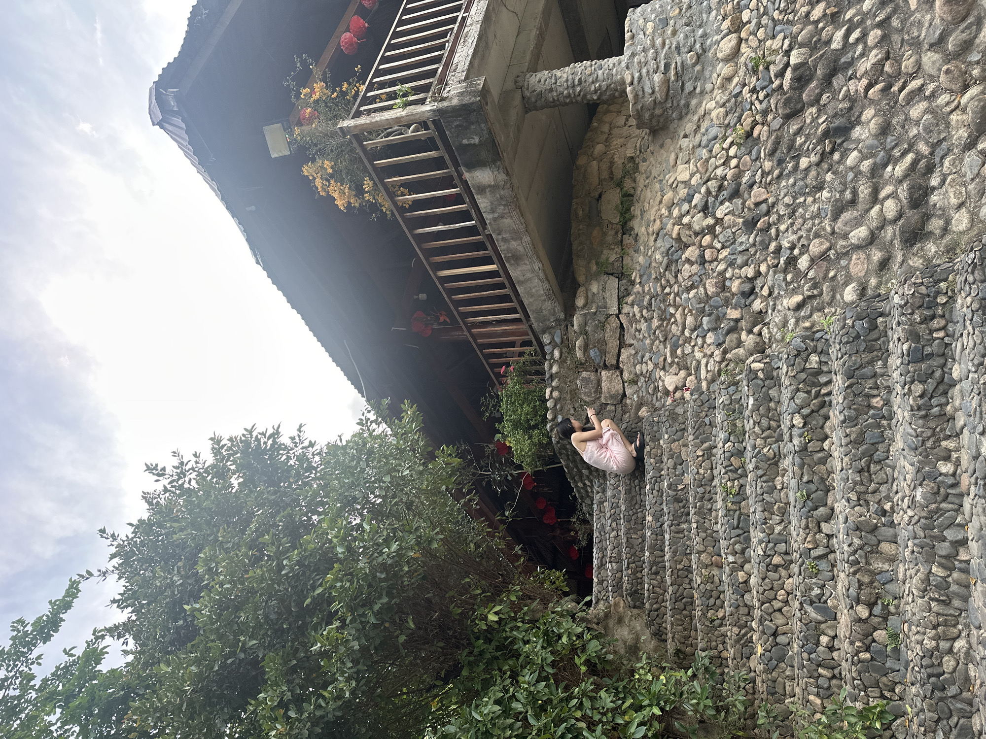 A low-angle, exterior shot depicts a woman sitting on stone steps in Nam Khanh Vinh, Vietnam. The steps, constructed of cobblestones, lead from the bottom right of the frame up to a building. The woman, wearing a light pink dress and dark sandals, sits with her back to the viewer, both hands raised to her head. The building is a weathered structure with a balcony, decorated with red lanterns and flowering plants. The building’s facade and support structures appear to be made of the same cobblestone material as the steps. A dense, green bush with small leaves is in the lower left of the frame, partially obscuring the lower portion of the building. The sky is overcast.