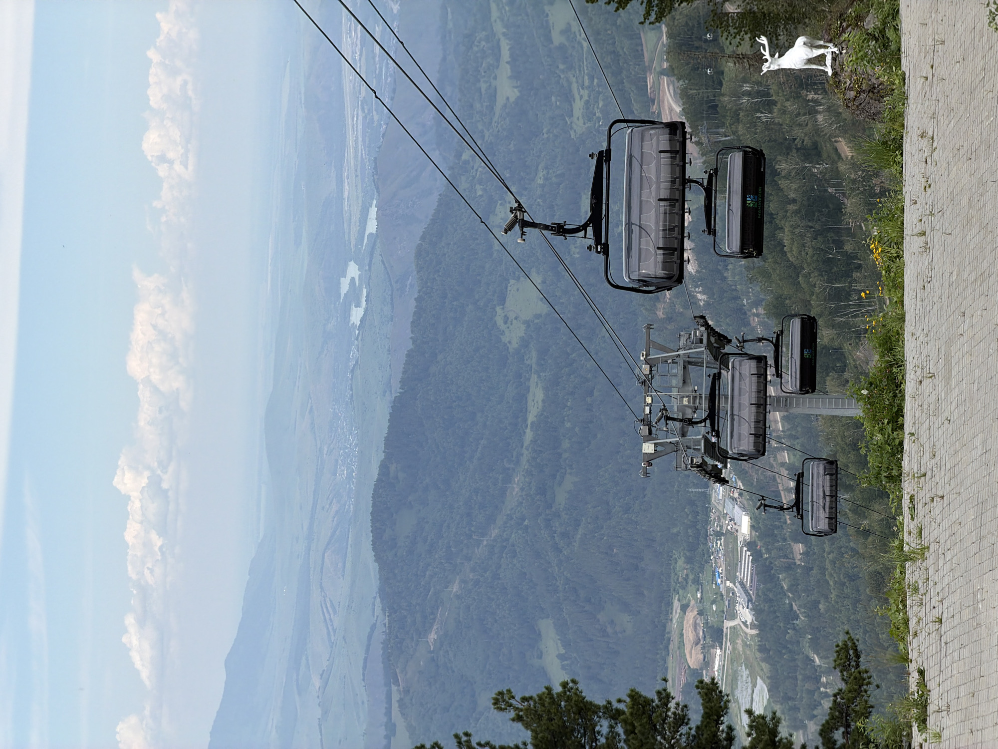 The image shows a scenic view from a ski resort with a cable car system in the foreground. Several cable car cabins are visible, suspended from steel cables that stretch across the frame. The cabins are dark and transparent, allowing a view of the empty seats inside. In the background, a vast expanse of rolling hills and mountains covered in lush green forests unfolds. A river winds through the valley, and a small settlement or town is visible in the distance. The sky is a pale blue with scattered white clouds. On a rocky outcrop to the right, a white statue of a deer stands prominently. The foreground features a paved pathway with scattered patches of grass and small yellow flowers. The overall scene suggests a tranquil and picturesque mountain landscape, likely at the Озерное ski resort in the Майминский district of Russia. There are no people visible in the image.