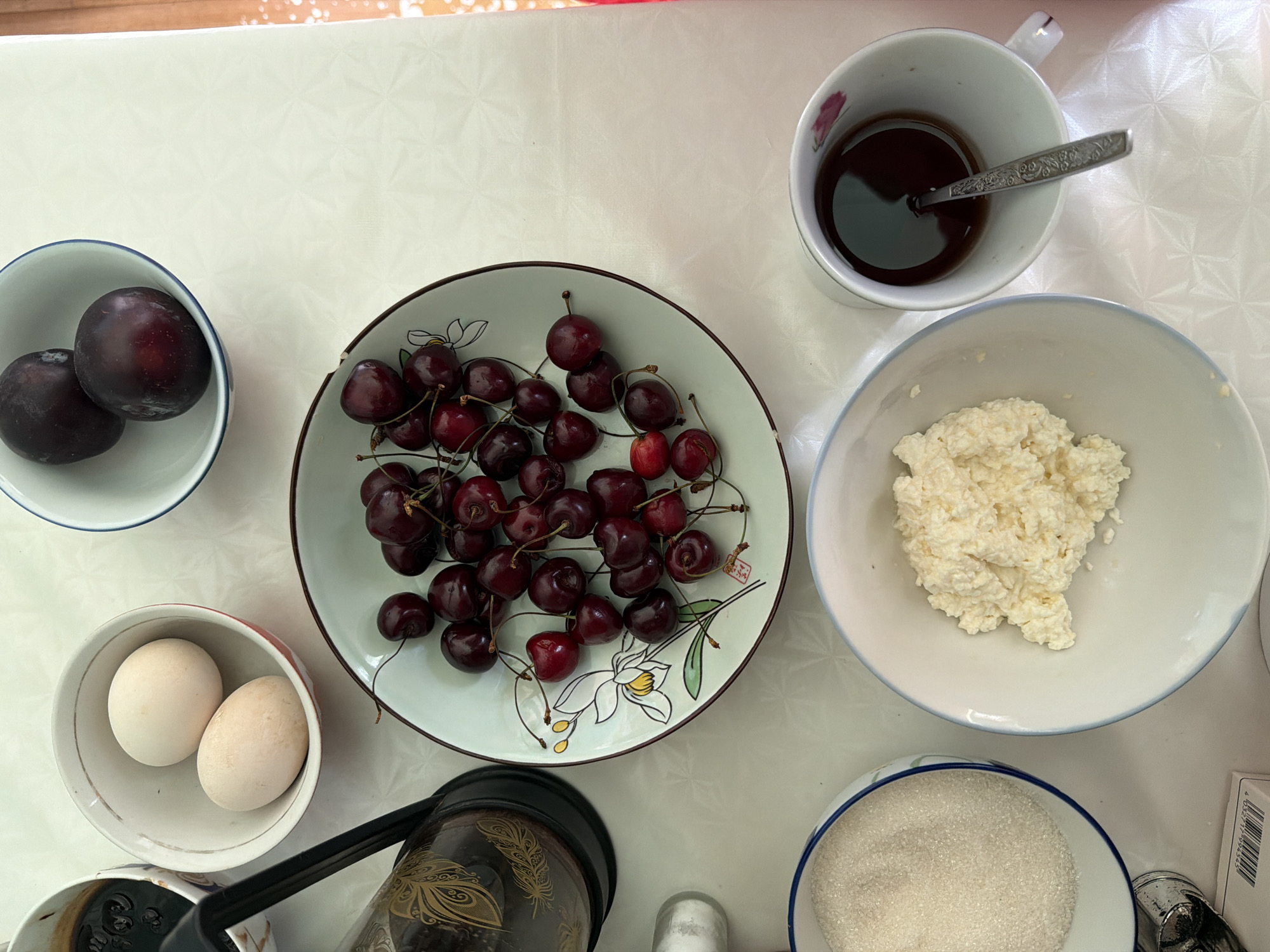 A top-down, eye-level shot captures a breakfast spread on a white, textured tablecloth. In the upper left is a dark brown French press with a black lid and handle, partially obscured by a small, white bowl containing two light brown eggs. To its right, a light blue and white rimmed bowl holds two dark purple plums.
The center of the image is dominated by a light green, circular plate with a floral design, filled with numerous dark red cherries. To the left of the plate, and slightly below, a white bowl with a blue rim contains white granulated sugar. To the right of the sugar bowl and below the cherry plate is a large, white, shallow bowl filled with a light yellow, clumpy substance, likely cottage cheese. In the bottom right corner, a white mug with a pink floral design contains dark brown liquid, presumably coffee or tea, with a silver spoon resting inside. The overall scene suggests a home setting, possibly a kitchen or dining area, during daylight.