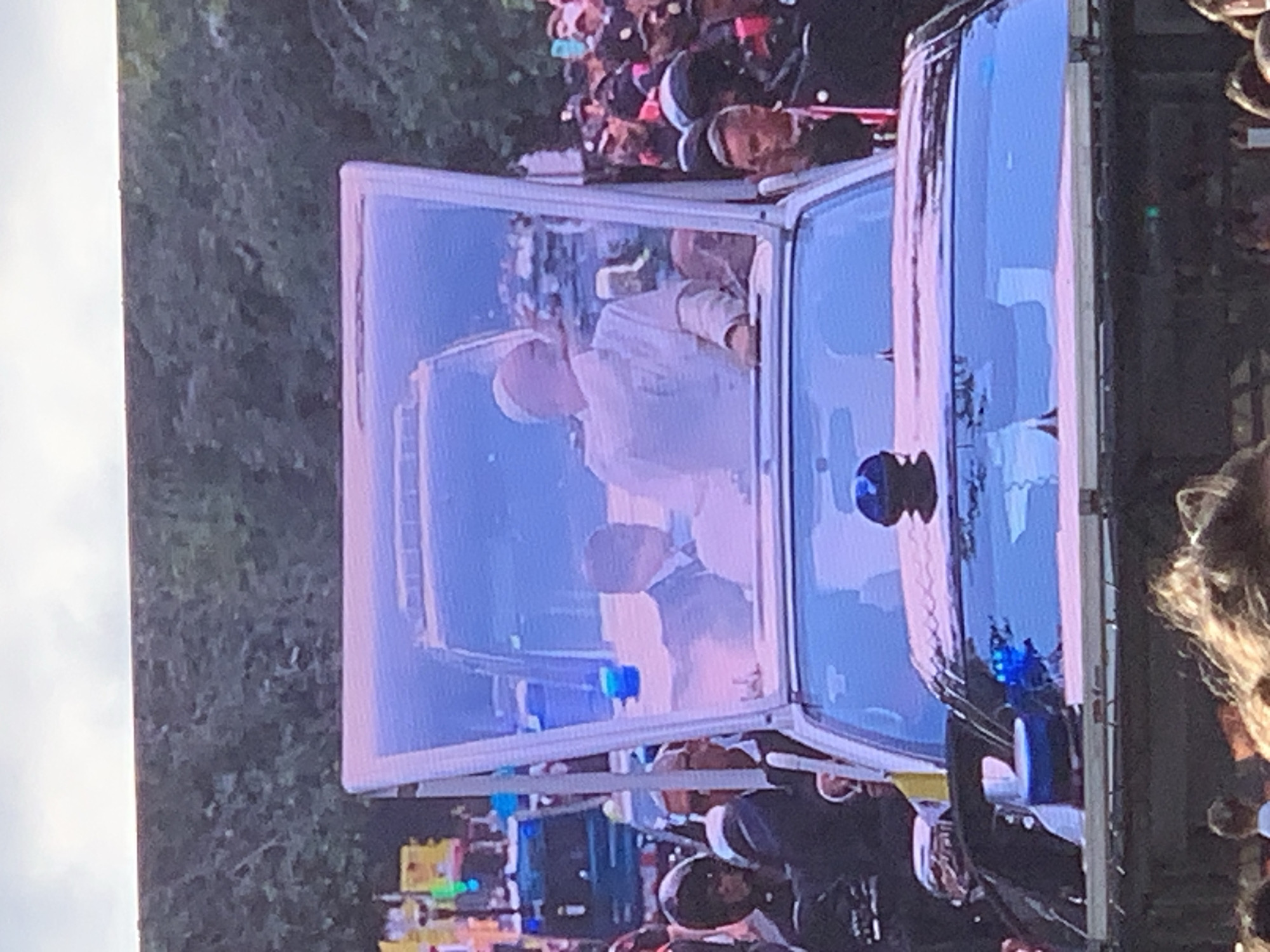 The image shows Pope Francis in a white papal cassock waving to a crowd. He is inside the popemobile, a vehicle designed to allow him to be visible and accessible to onlookers. The popemobile has large glass windows, offering clear views of the Pope. The scene appears to be outdoors, possibly during a public appearance or procession. The background is blurred, but hints of green foliage suggest a park or an area with trees.
The Pope's demeanor appears friendly and engaged as he waves. He is the central subject, his white attire making him stand out against the blue tones of the popemobile and the surrounding environment. A blue light is visible on the popemobile, possibly indicating a safety or official function. In the crowd, people are visible, suggesting a significant gathering. Some individuals are holding up their phones, likely to capture the moment. The overall mood seems to be one of public admiration and religious significance. There is no visible text in the image. The time of day is not explicitly clear, but the lighting suggests it could be daytime.