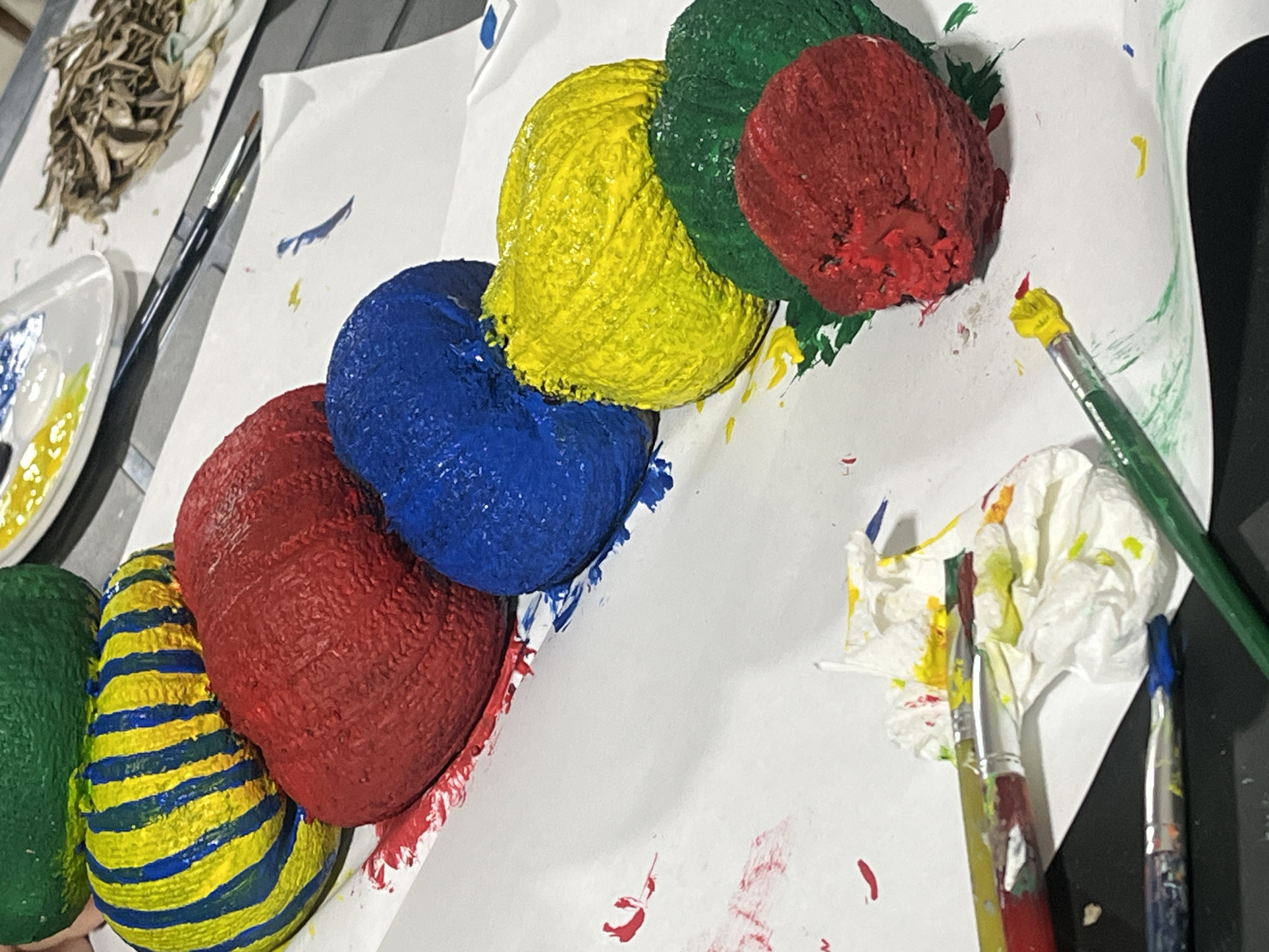 The image depicts a close-up, high-angle shot of several round, painted objects, likely rocks or gourds, arranged diagonally on a white paper surface. The objects are painted in bright, solid colors and patterns: a green object is at the top, followed by a yellow object with blue stripes, then a red object, a blue object, a yellow object, a green object, and finally a red object at the bottom. Paint splatters in red, yellow, blue, and green are visible on the paper around the objects. Several paintbrushes are also present, some with paint on their bristles, and a crumpled white paper towel with paint stains lies near the center. The background is blurred, but appears to be a dark, flat surface, possibly a table. There is no indication of the city of Surami, Georgia, or any people in the image.