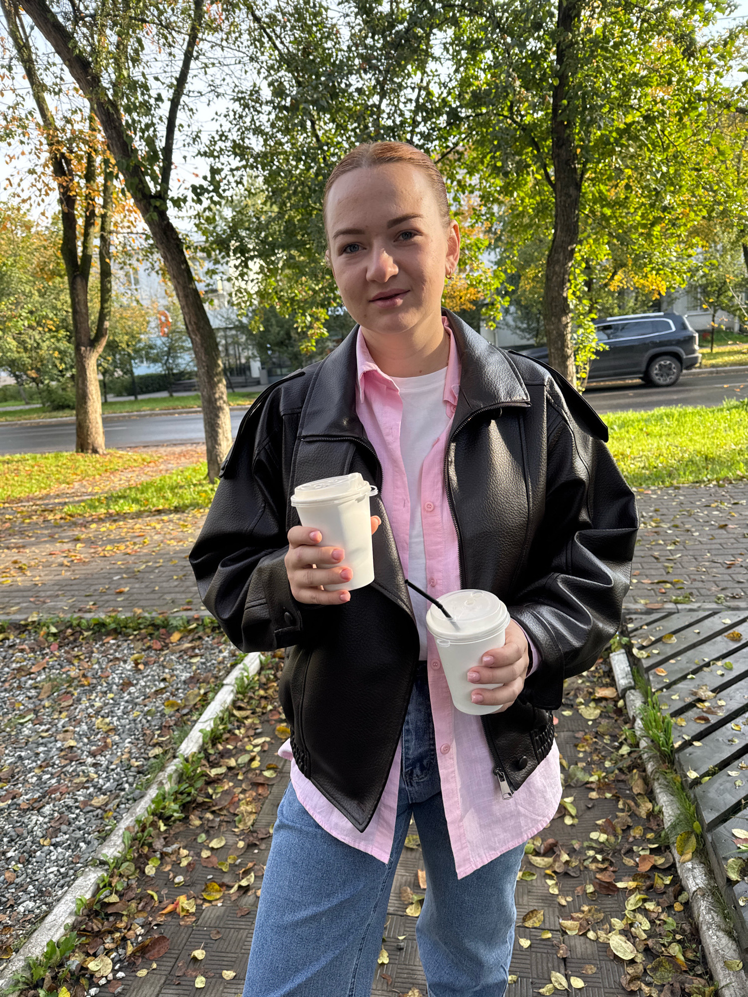 A woman stands outside, holding two white paper cups with lids, one with a straw. She is wearing a black leather jacket over a pink collared shirt and blue jeans. The background shows trees with autumn leaves, a sidewalk, a street, and a parked car. The scene appears to be in a park or residential area of Chaikovsky, Russia, judging by the architecture of the building visible in the distance. The lighting suggests it is daytime.