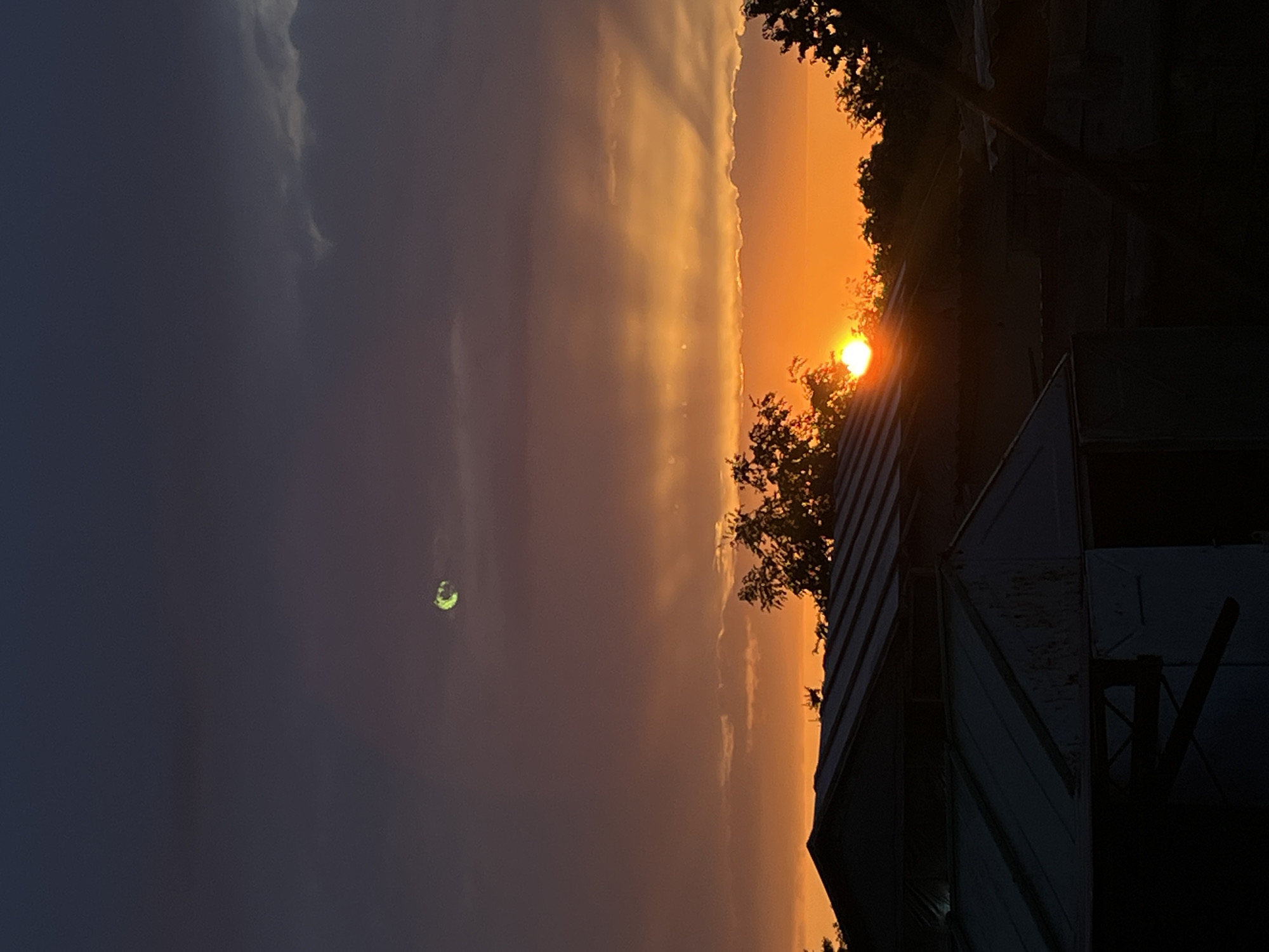 The image captures a sunset over a rural or suburban area. Silhouetted against the vibrant orange and purple sky are rooftops of buildings and the foliage of trees. The sun is partially visible, casting a bright glow on the horizon. In the upper portion of the sky, a faint, greenish-tinged object, possibly the moon or a lens flare, is discernible. The scene suggests the end of the day in a quiet, undeveloped location.