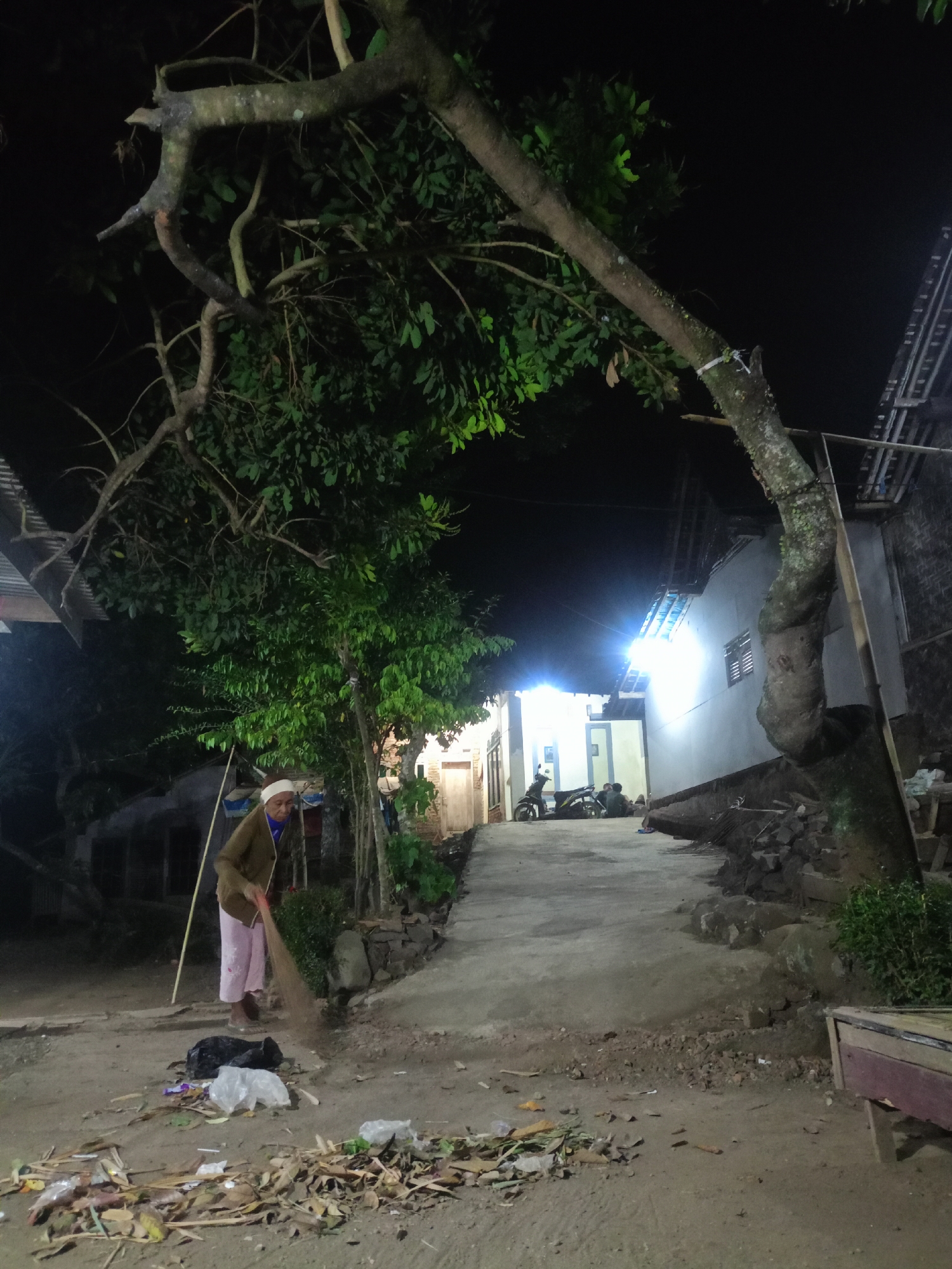 In the village of Jelbuk, Indonesia, at night, a woman wearing a white head covering and a brown jacket over pink pants sweeps debris on a dirt path. The path is lined with scattered leaves, twigs, and plastic waste. To the left, a large tree with twisted branches overhangs the scene. In the background, several houses are visible, some illuminated by bright lights, and two motorcycles are parked near one of the buildings. A pile of rocks and construction materials sits near the base of a large tree on the right.