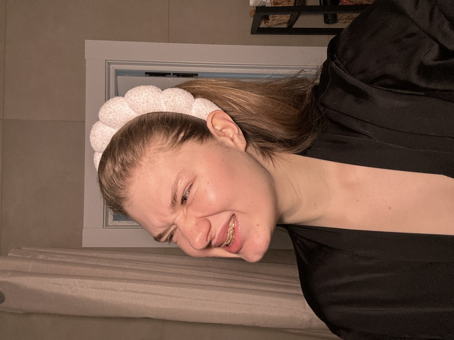 The image depicts an indoor scene in the city of Nikol'skii, Russia. A light-skinned female individual, with blonde hair pulled back into a ponytail, is positioned centrally. She wears a white, puffy-textured headband and a black, satin-like robe. Her facial expression is a pronounced grimace, characterized by tightly squinted eyes, a deeply wrinkled nose, and an open mouth revealing gold-colored dental braces on her upper teeth. To the left of the subject, a light-colored vertical curtain is visible. Behind her, a white doorframe with molding is present. To the right, a dark, multi-shelf storage unit is partially visible, containing several light-colored objects, possibly baskets or containers. The scene is illuminated by warm, artificial light.