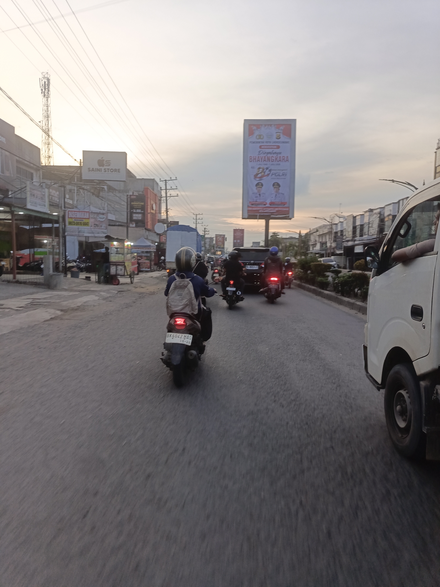 The image depicts a street scene in Lhokseumawe, Indonesia, likely during the late afternoon or early evening due to the soft, diffused lighting and the sky's gradient. The perspective is from a vehicle moving along a paved road.
In the foreground, the rear of a white truck is visible on the right side of the frame. A motorcycle with a rider wearing a helmet and a beige backpack is in the center, traveling in the same direction as the truck. The motorcycle's license plate reads "BK 6042 MAO 10-28." Behind this motorcycle, several other motorcycles are also on the road, some with riders.
The street is lined with buildings on both sides. On the left, there are shops, including one with a prominent sign that reads "SAINI STORE." Further down the road, a tall billboard displays graphics and text, including an image of two men in official attire and the words "PEMERINTAH KOTA LHOKSEUMAWE" and "BHAYANGKARA." On the right side of the road, there are more buildings and some greenery.
Overhead, a network of power lines crisscrosses the sky, and a telecommunications tower is visible in the distance on the left. The sky is mostly cloudy with a soft, warm light, suggesting either sunrise or sunset. Traffic appears moderate, with vehicles and motorcycles sharing the road. The overall impression is a typical urban street scene in Indonesia, bustling with activity.