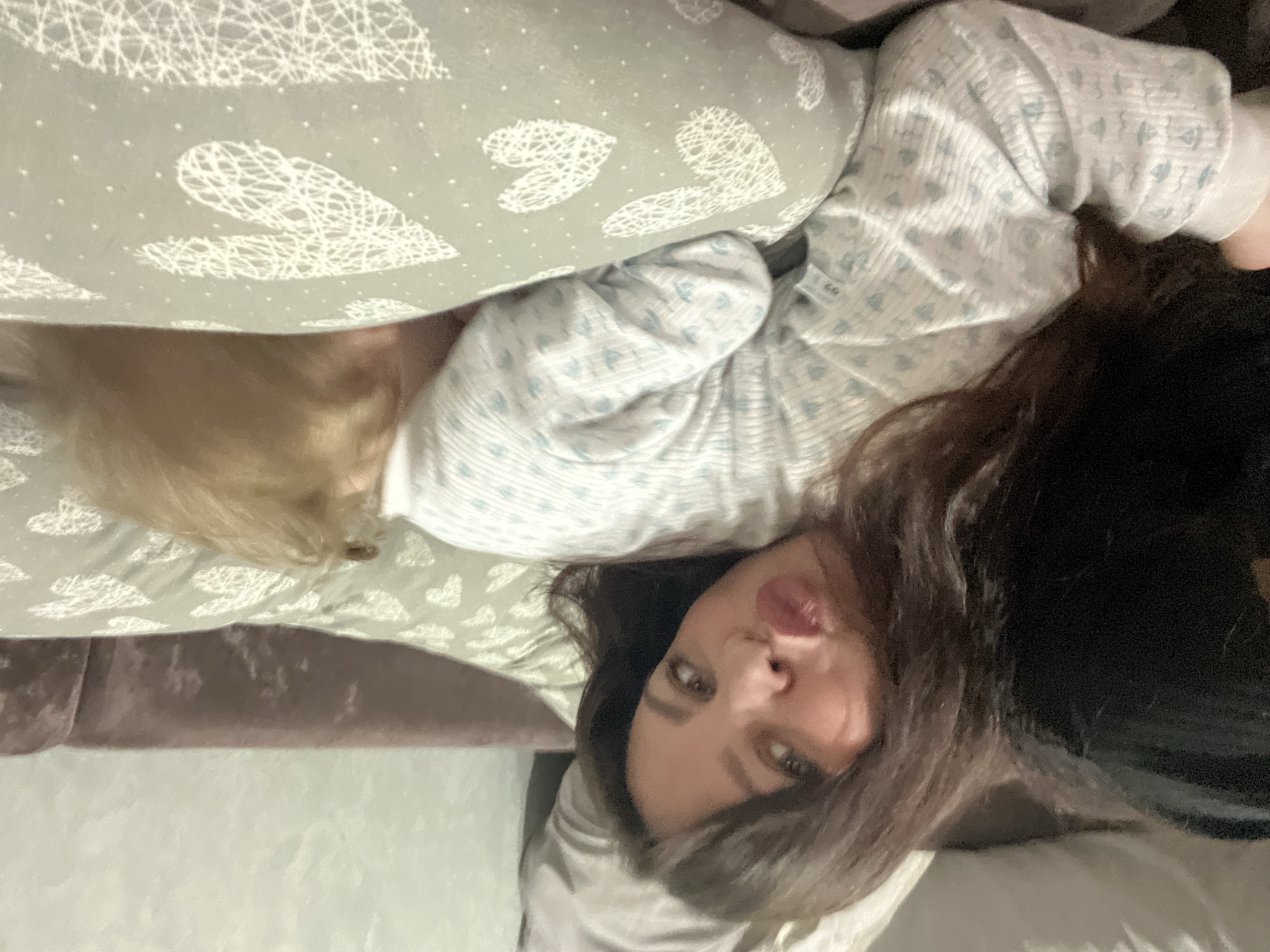 A woman is lying on a bed with a pillow patterned with white hearts. She has dark brown hair and is wearing a light blue and white patterned shirt. Part of a blonde-haired person is also visible on the bed. The environment suggests an interior setting, possibly a bedroom. The location is not evident from the image.
