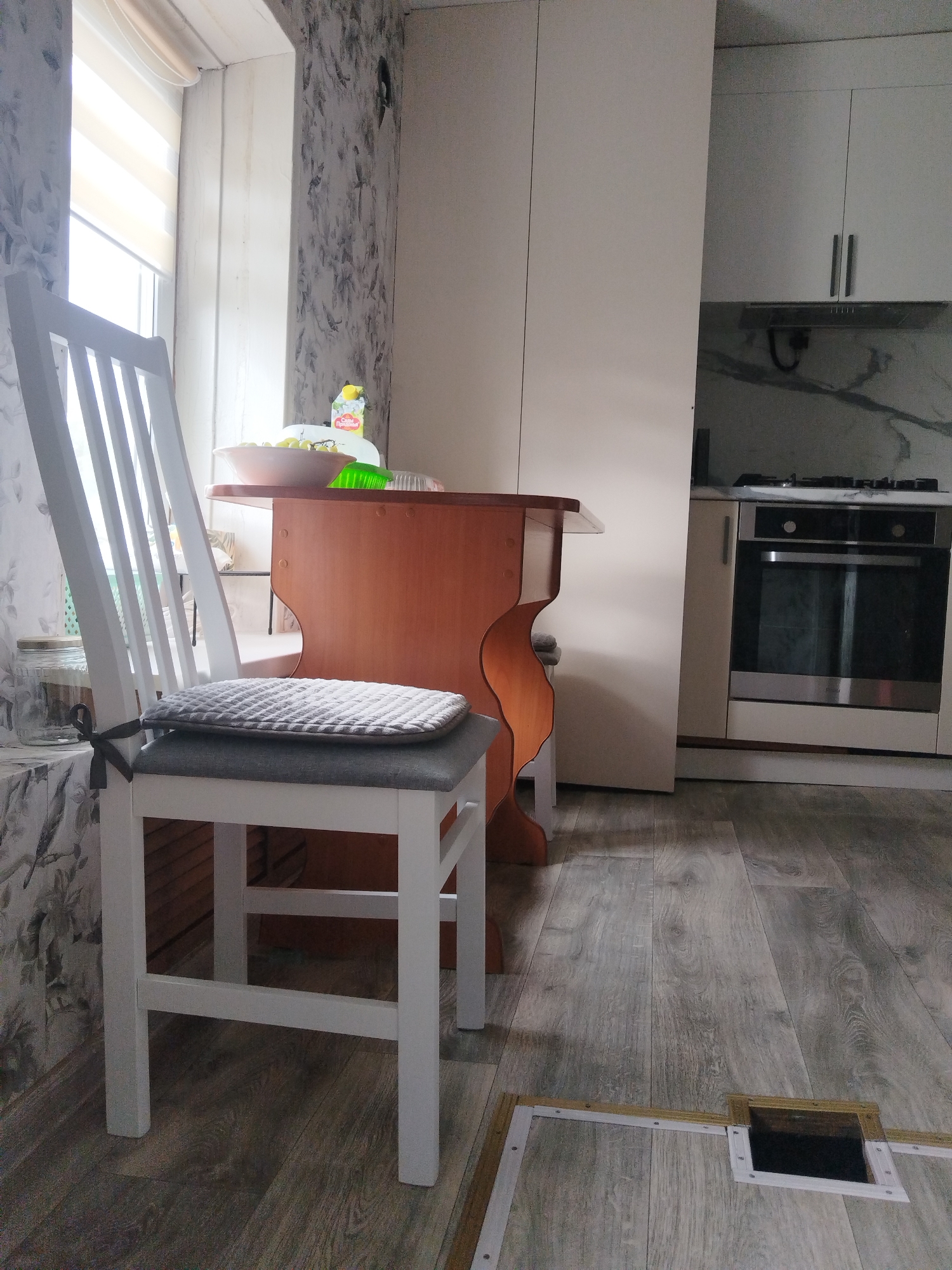 The image depicts an indoor scene, likely a kitchen or dining area, with a white wooden chair in the foreground. The chair has a grey upholstered seat and a quilted grey cushion tied to its back. To the right of the chair, a wooden table with a curved base is visible, holding a bowl of grapes and a bottle of liquid. In the background, kitchen cabinets and a stainless steel oven are present. The floor is covered with grey wooden laminate flooring, and a square opening with a white frame is visible on the floor in the lower right portion of the image. The walls are decorated with floral wallpaper. The overall impression is that of a domestic setting. There are no individuals or specific actions depicted in the image. The location within the city of Votkinsk, Russia, cannot be determined from the visual information provided in the image.