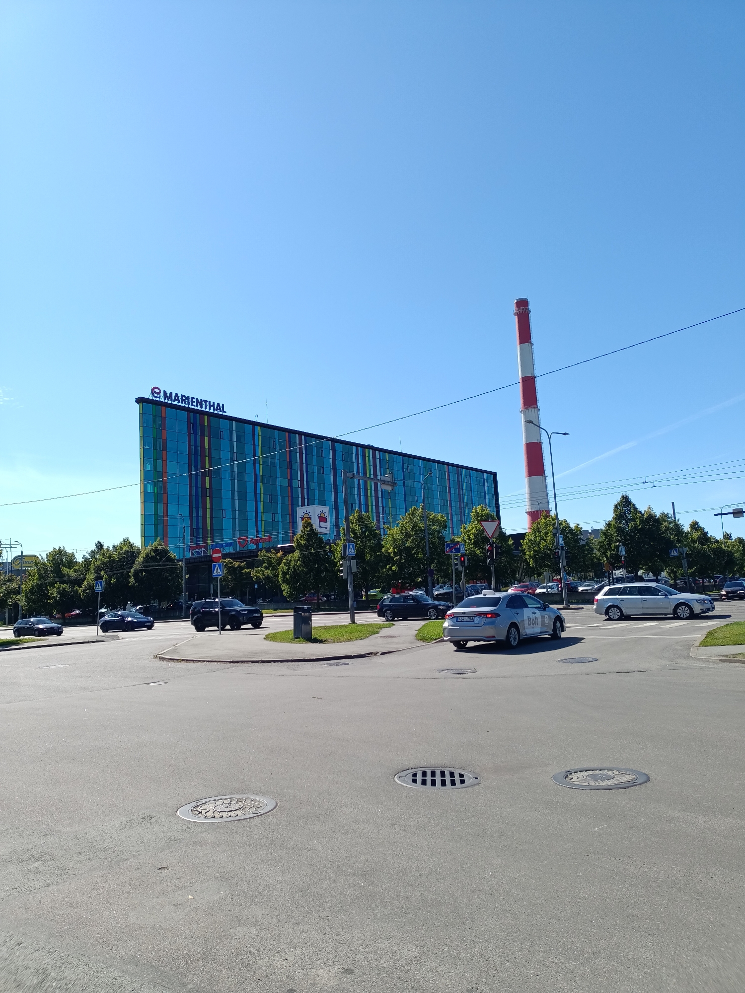 A bright, sunny day in Tallinn, Estonia, is depicted in this image, which captures a street scene with a modern, colorful building and a prominent red and white striped smokestack in the background. The building, labeled "MARIENTHAL," features a glass facade with vertical strips of various colors. Trees line the street, and the sky is a clear, vivid blue, suggesting fair weather.
The foreground shows a paved intersection with several cars navigating the road. Among them are a silver sedan with "Bolt" branding on its side, a dark SUV, and other vehicles. Traffic appears to be moving steadily. Several manhole covers are visible on the asphalt, indicating urban infrastructure.
The red and white smokestack stands tall, a distinct industrial feature against the cityscape. The overall impression is one of a functional urban environment on a pleasant day. No people are clearly discernible in the image. The image was likely taken in the daytime due to the strong sunlight and clear visibility.