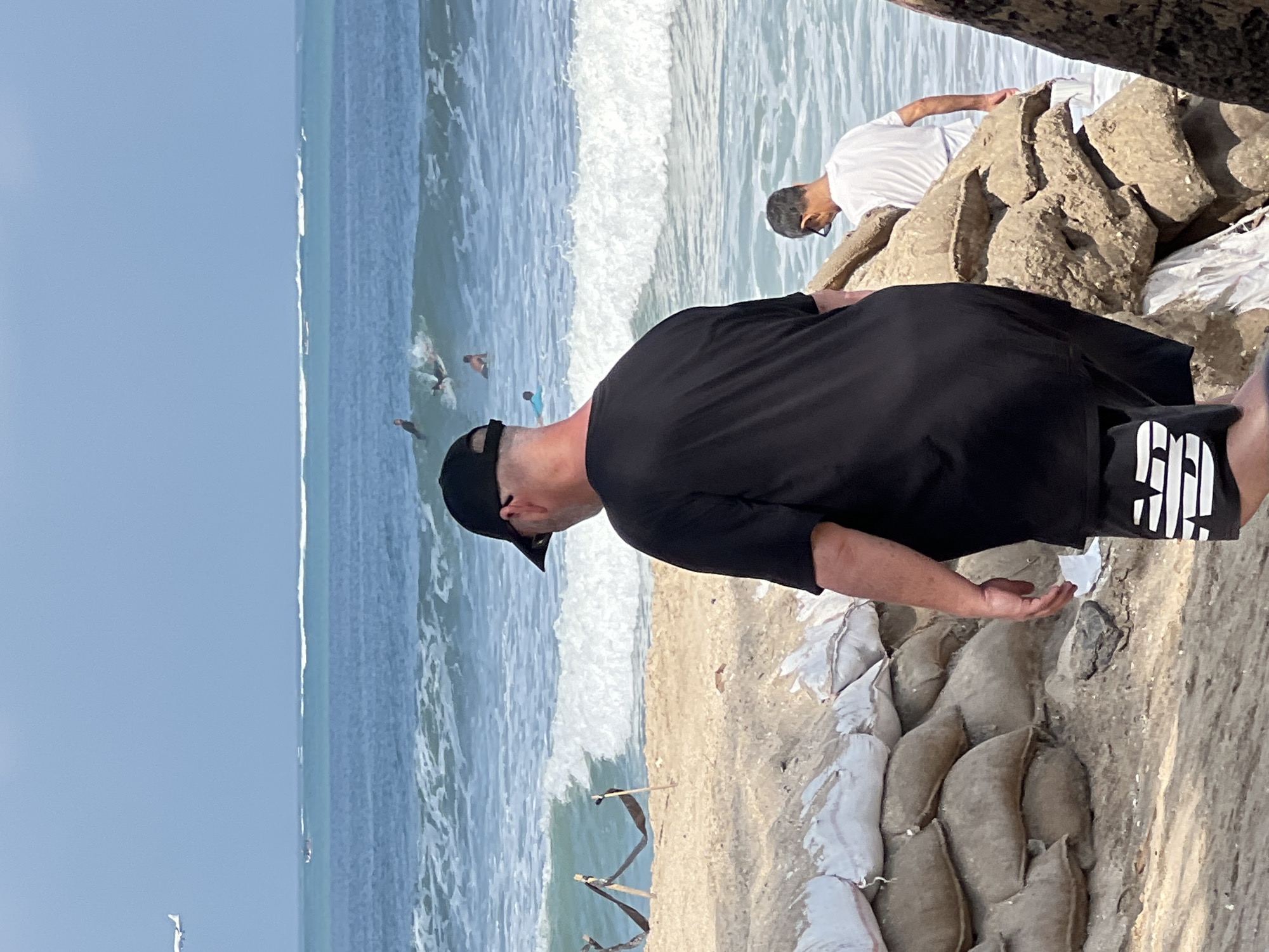An adult male wearing a black t-shirt, black shorts with a white logo, and a black baseball cap is walking away from the camera towards the ocean on a sandy beach. In the background, several surfers are riding waves. Another adult male wearing a white t-shirt and sunglasses is standing in the shallow water. Sandbags are stacked along the shoreline, presumably for flood protection. An airplane is visible in the sky. The environment is a beach with a blue ocean and clear sky. The location is Kuta, Indonesia.