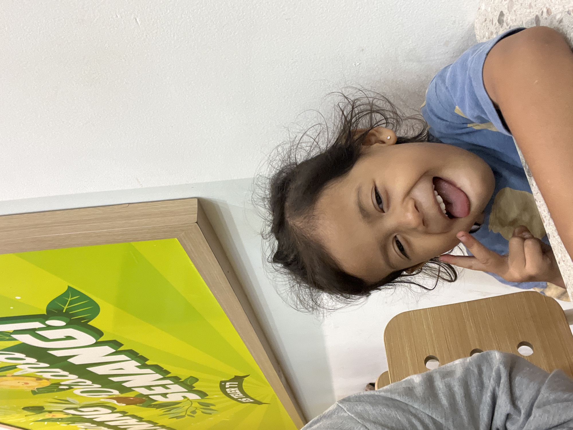 A young girl with dark hair, wearing a blue shirt, is pictured indoors. She is sticking her tongue out and making a peace sign with her fingers. Behind her, a brightly colored sign with green lettering that appears to read "SENANG!" and "ES TELER 77" is visible, framed by a light brown border. The wall behind her is white and textured. Part of a wooden chair and a person in a grey shirt are also visible to the left. The scene suggests the girl is at a food establishment, possibly a restaurant or cafe, in the city of Gunung Putri, Indonesia.