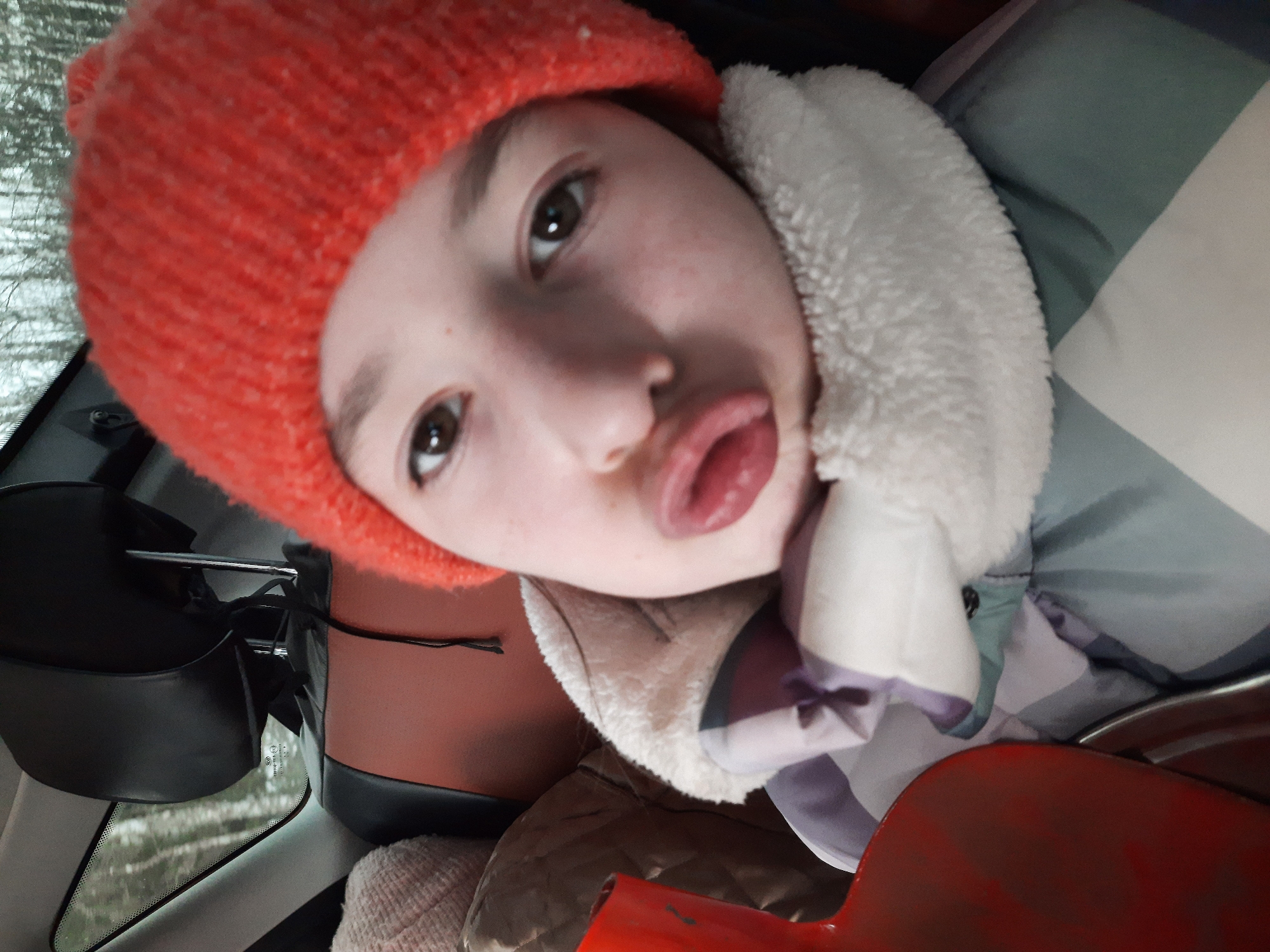 A person wearing a red knit hat and a white faux fur-lined jacket is taking a selfie in what appears to be the interior of a car. The person has light skin, brown eyes, and is sticking their tongue out. A car headrest is visible in the background along with the windshield showing some bare trees. The location of the image is Архангельское, Russia, as specified in the request. Other objects present include an interior car storage pocket, the car’s windshield, and an unidentified red item close to the bottom of the frame.