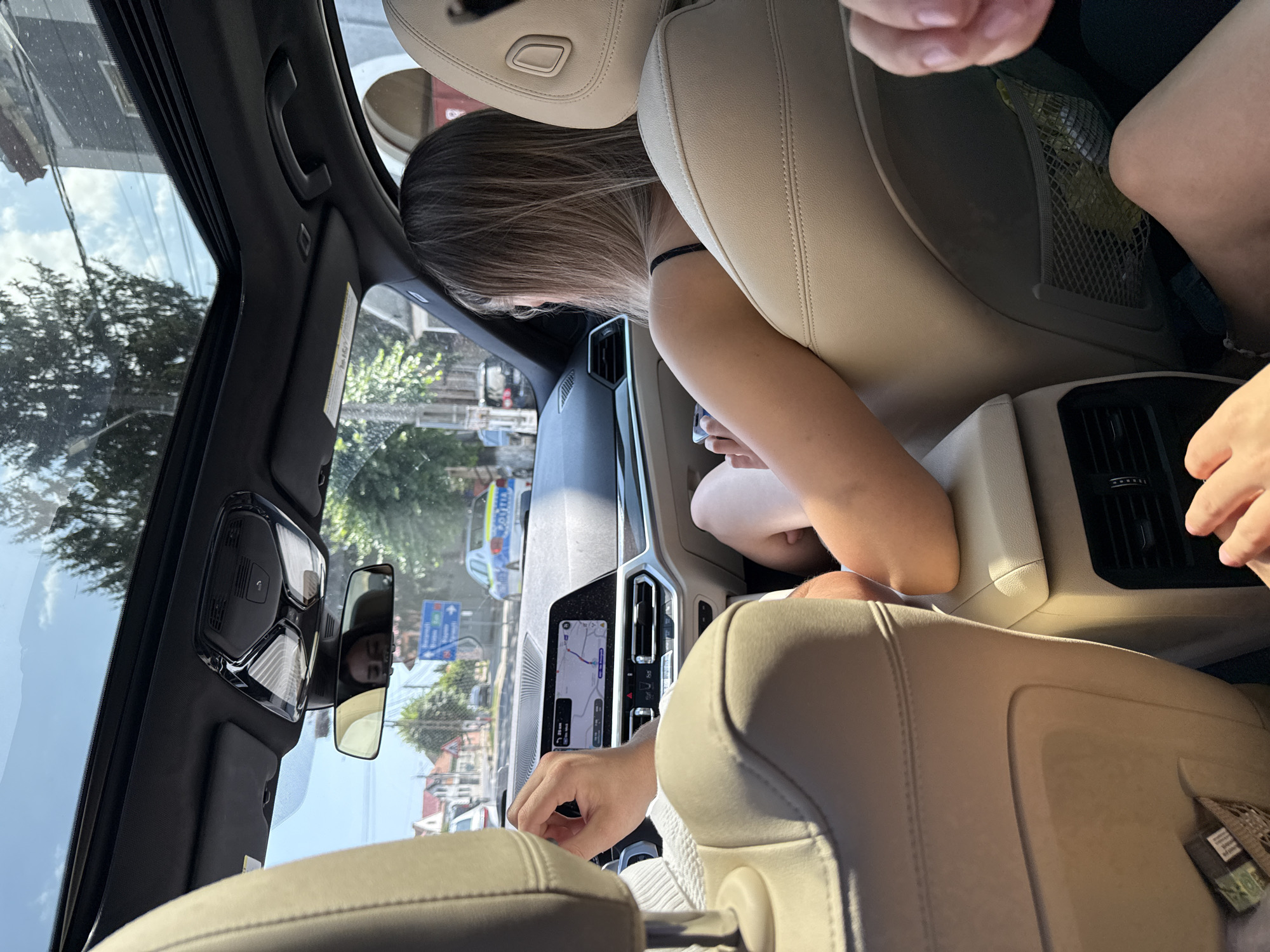 The image, taken from the interior of a car, shows a view out of the windshield and side windows, suggesting the car is in motion. The scene depicts a street in Șercaia, Romania. A police car with visible markings is present on the road, indicating a potential law enforcement activity or presence. The interior of the car includes the dashboard with a navigation screen, a rearview mirror reflecting a person's face, and beige leather seats. Two individuals are partially visible within the car: one in the driver's seat with their hand on the steering wheel, and another in the passenger seat, looking out the window. The environment outside the car features buildings, trees, and overhead power lines, consistent with a typical urban or semi-urban setting.