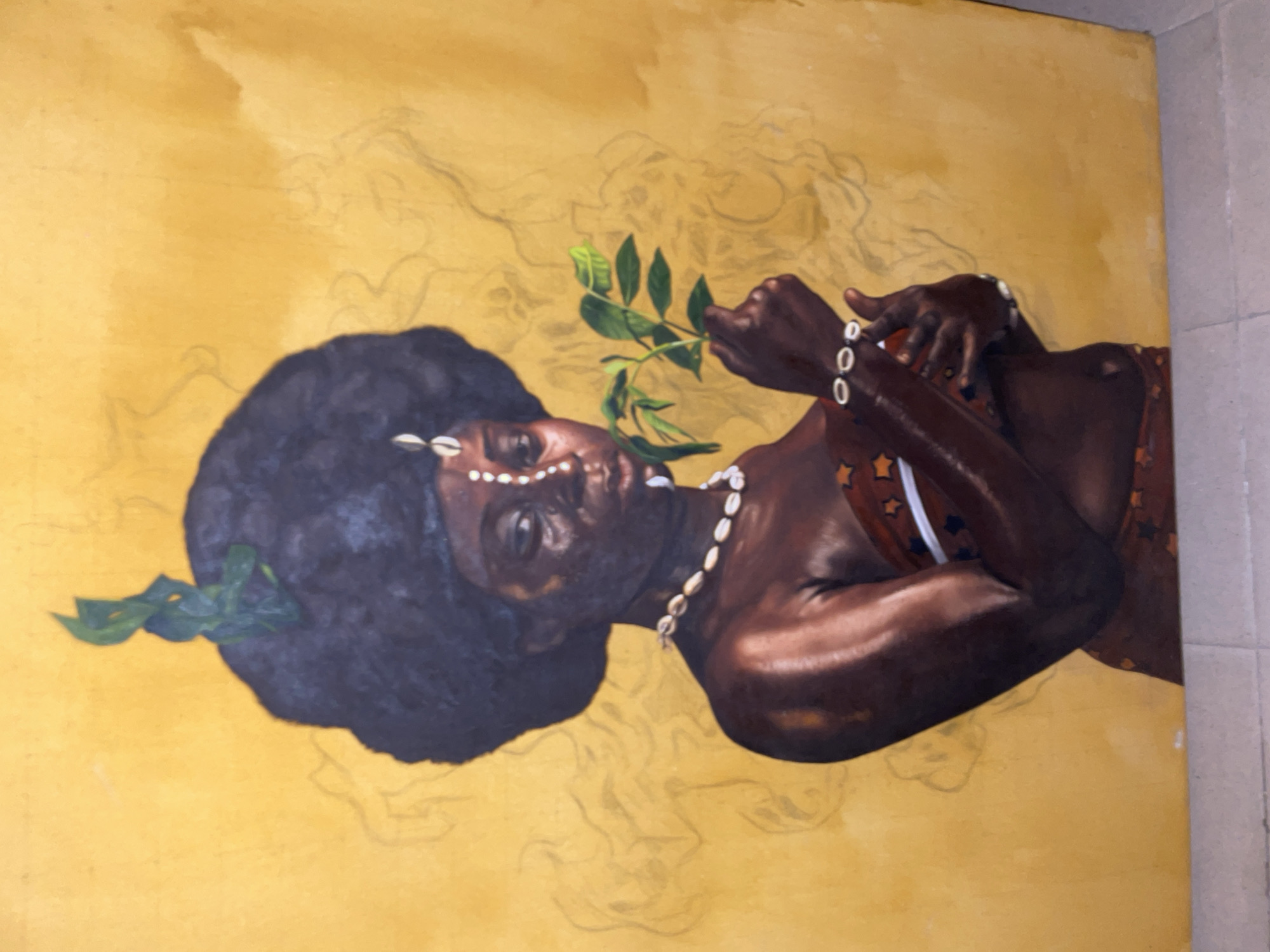 The image displays a painting, captured in a rotated landscape view, depicting a dark-skinned individual, likely female, with a prominent afro hairstyle. The subject's skin tone is rich brown with highlights suggesting muscle definition on the arms and shoulders. She wears adornments made of cowrie shells, visible as a necklace around her neck, decorative elements on her forehead, and bracelets on her wrists. One hand holds a vibrant green leafy branch, which contrasts against her dark skin and hair. She also appears to be wearing a top with a star pattern and white trim.
The background of the painting is a warm, golden-yellow hue, which covers most of the surface. Faint, ethereal pencil or charcoal outlines of swirling, organic shapes are visible across the yellow background, especially around the subject's head and the upper right portion of the painting. These outlines suggest either an incomplete stage of the painting process or a deliberate artistic choice to create an abstract or ethereal atmosphere. The subject's gaze is directed towards the viewer, with an expression that appears serene and contemplative. The overall aesthetic, including the subject's features and use of cowrie shells, aligns with cultural representations often found in African art, consistent with the location context of Ado, Nigeria. There is no visible text in the image.