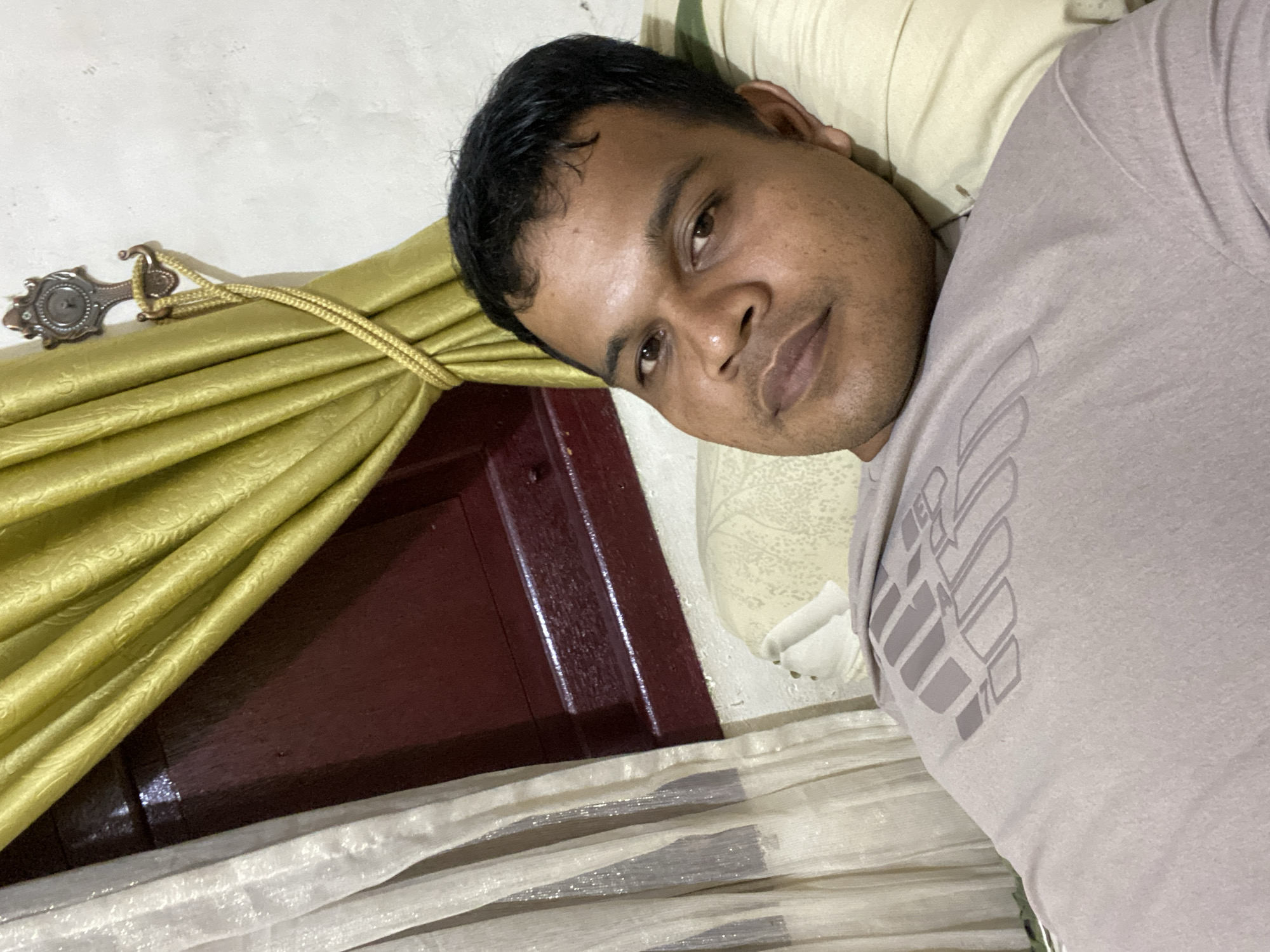 A man lies on a bed, his head resting on a pillow. He is wearing a light purple t-shirt with a grey graphic design. To his left, a sheer white curtain with metallic threads is visible, and behind him, golden curtains are tied back with a rope to a decorative hook. The background appears to be a wall. The man's facial expression is neutral, and he is looking slightly away from the camera. The lighting suggests an indoor setting at night.