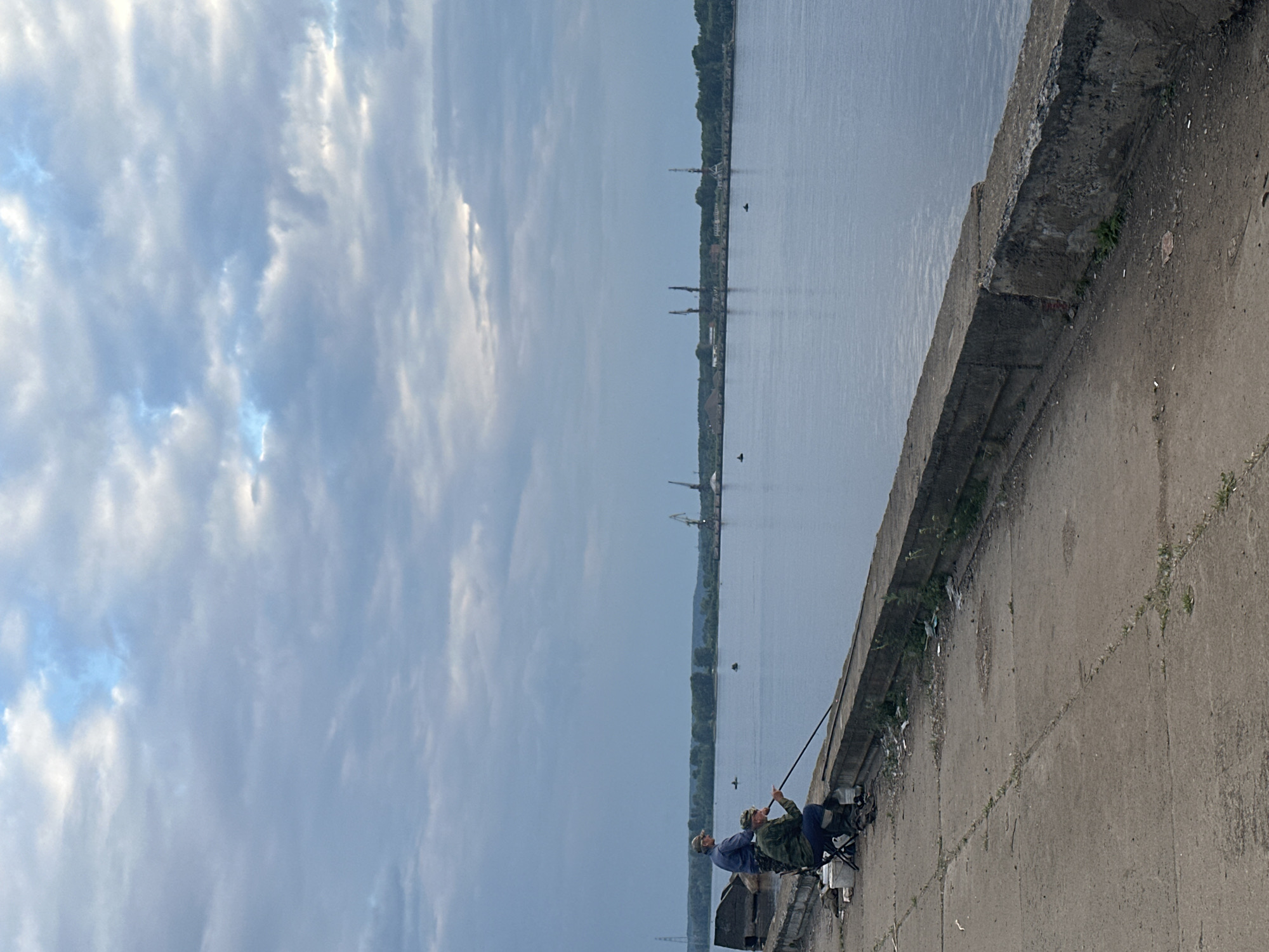 The image depicts two individuals fishing from a concrete embankment beside a wide river. The sky is overcast with scattered clouds. On the left side of the embankment, one person is seated on a black folding chair, holding a fishing rod and seemingly smoking a pipe. Beside them is a white bucket. Another person stands to the left of the seated individual, also with a fishing rod. The embankment is made of weathered concrete, with some debris and sparse vegetation growing in cracks. In the distance across the river, there is a treeline, and further beyond, several industrial cranes are visible. The water is calm, reflecting the cloudy sky.