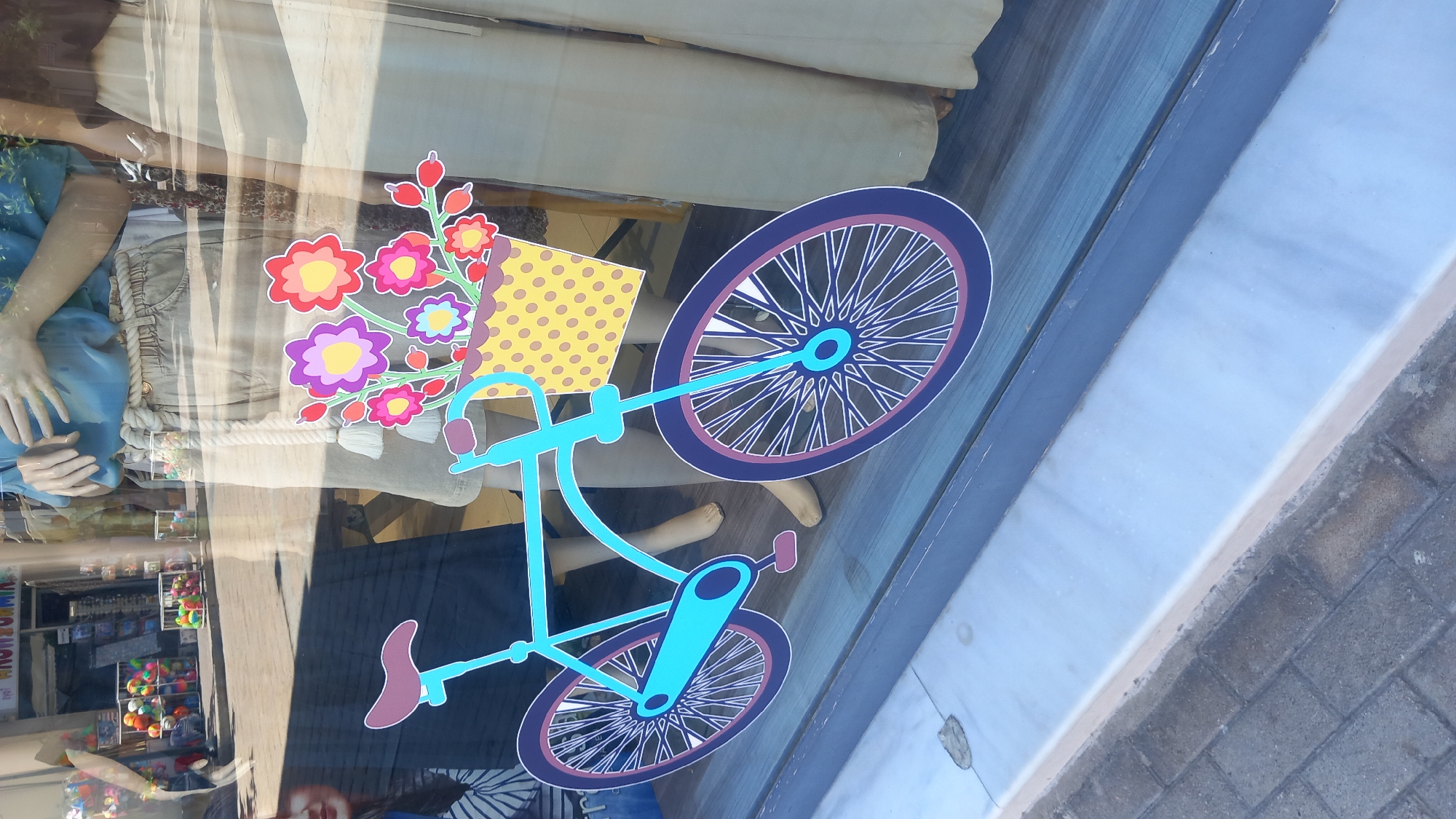 The image shows a close-up of a shop window in the city of Kos, Greece, with reflections of the street and surrounding buildings visible. A stylized, cut-out image of a turquoise bicycle with purple and pink accents is adhered to the glass. The bicycle has a basket filled with colorful flowers. Mannequins are visible behind the window display, dressed in casual clothing. The ground outside the shop is paved with grey cobblestones. The window is set within a blue frame, and the sill appears to be made of white marble. The overall impression is of a cheerful, summery display.