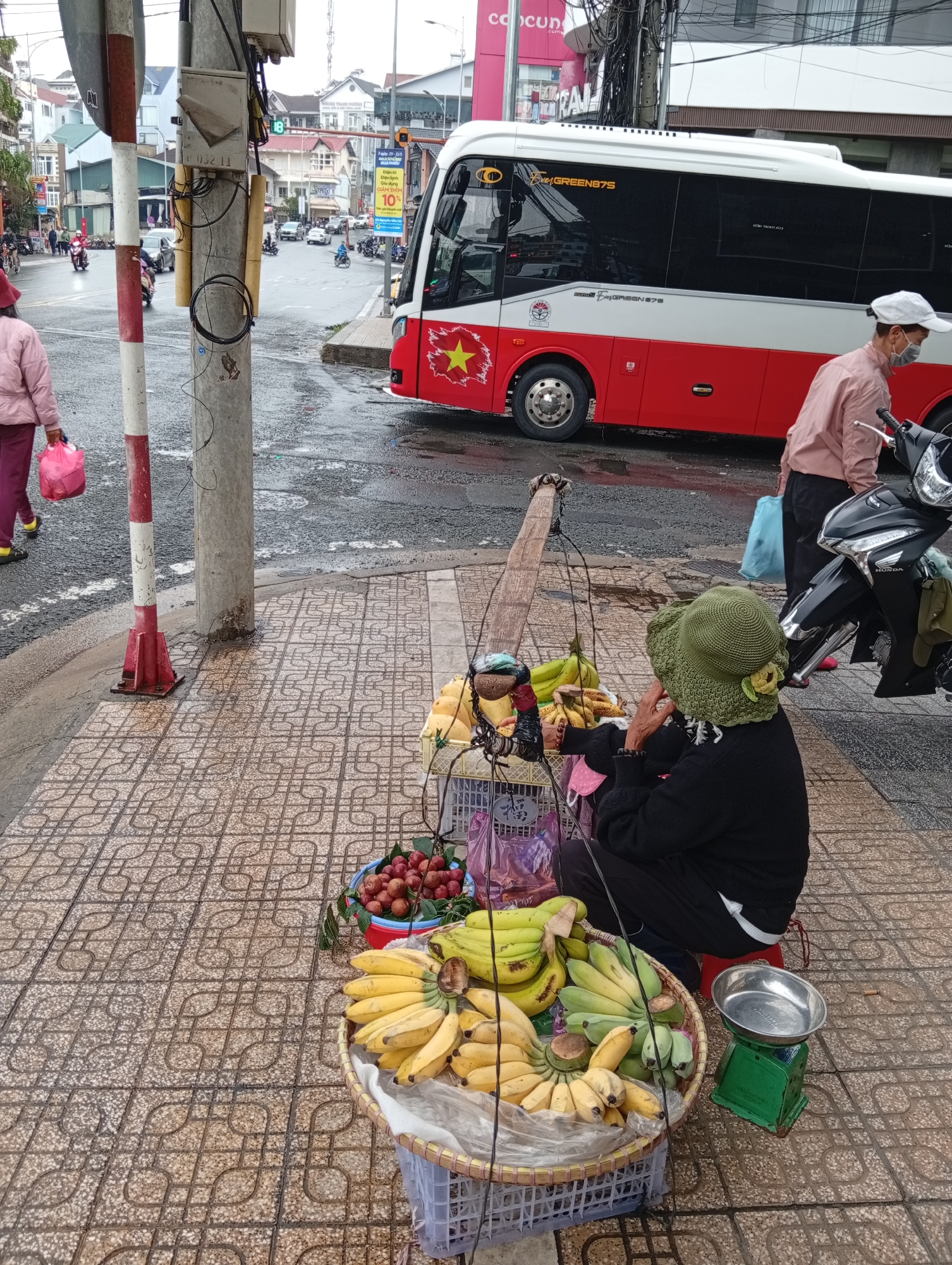 Captured from a high-angle perspective in the city of Da Lat, Vietnam, the photograph depicts a street scene where a female fruit vendor is seated on a low red stool on a paved sidewalk. The vendor is clad in a black top and trousers, and her head is adorned with a green crocheted hat. She is positioned behind two baskets overflowing with bananas, complemented by a smaller basket filled with lychees or similar fruits. Next to her is a green and silver analog scale for weighing produce.
A traditional carrying pole, known as a "gánh," rests over her shoulder, laden with additional fruits. Behind the vendor, a man wearing a pink shirt, dark pants, and a white cap stands near a parked black Honda motorcycle.
Adjacent to the sidewalk is a wet street traversed by a red and white bus bearing a yellow star, possibly a Vietnamese bus service, with the words "EverGREEN875" on its side. Further down the street, there are additional vehicles, pedestrians, and buildings under an overcast sky. A street sign with Vietnamese writing and a phone box add to the urban context.