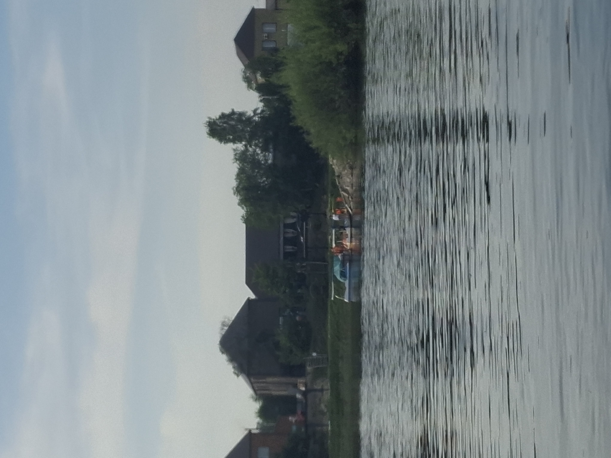 This is an outdoor, medium-range, slightly blurry, eye-level, full shot of a body of water with a shoreline and buildings. The water is rippled. The sky is mostly clear with some light clouds. On the shoreline are several buildings, some with dark roofs and windows. Trees are visible behind the buildings, some dense and green, others sparser. Two small boats are docked near the shore. One boat is blue and white with a cover, and the other appears to be orange and white, with several people on board wearing orange life vests. The scene is likely a residential or recreational area.