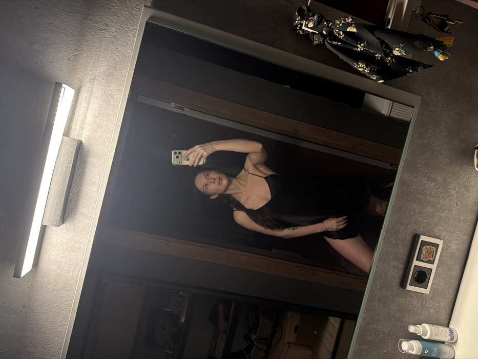 A bathroom mirror reflects a woman in a black slip dress holding a phone, presumably taking a picture. The reflection reveals a doorway behind her. A rectangular vanity light illuminates the left side. A black floral hair accessory hangs above the mirror. A rectangular box containing two small round holes and bottles of lotion are visible on the right counter. The image is taken at a slightly skewed angle.