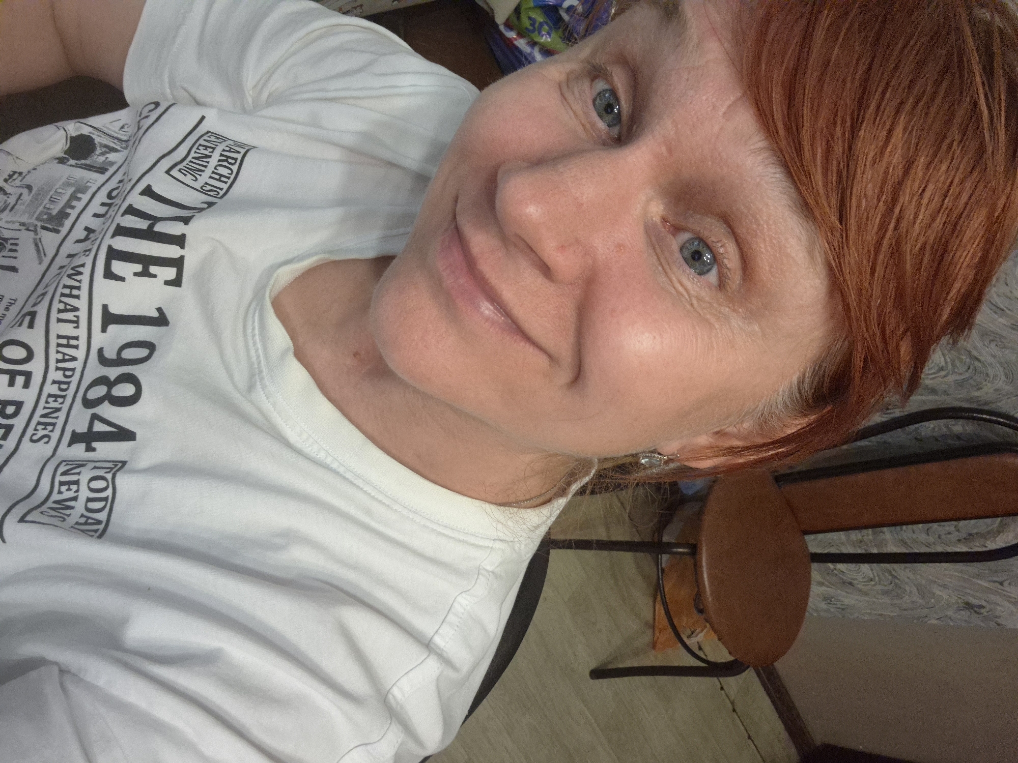 This media file is a selfie of a woman with reddish-blonde hair and blue eyes. She is wearing a white t-shirt with black text that appears to be part of a newspaper or magazine layout. The visible text includes "THE 1984", "WHAT HAPPENES", "OF", and "TODAY NEWS". The woman is looking at the camera with a slight smile. The background is out of focus but suggests an indoor setting, possibly a home, with hints of furniture and patterned wallpaper. The lighting appears to be artificial, suggesting it was taken indoors at any time of day. The overall mood of the image is casual and friendly. The selfie is taken from a close-up perspective, with the woman's face and upper body being the main focus. The orientation of the image is tilted.