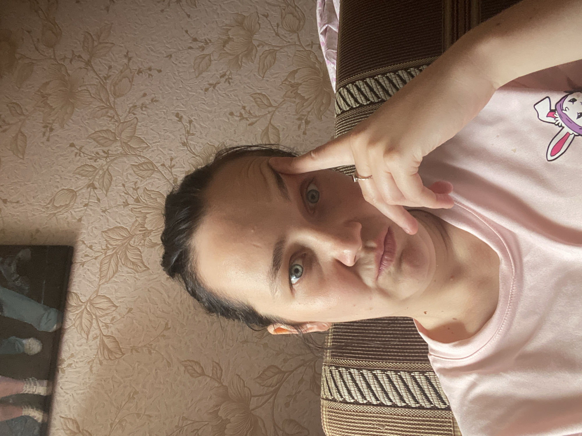 A woman is sitting on a couch and looking at the camera. She has her right hand on her forehead and a ring on her left ring finger. She is wearing a pink t-shirt with a cartoon bunny graphic. The background is a wallpaper with a floral pattern and a framed picture on the wall behind her.