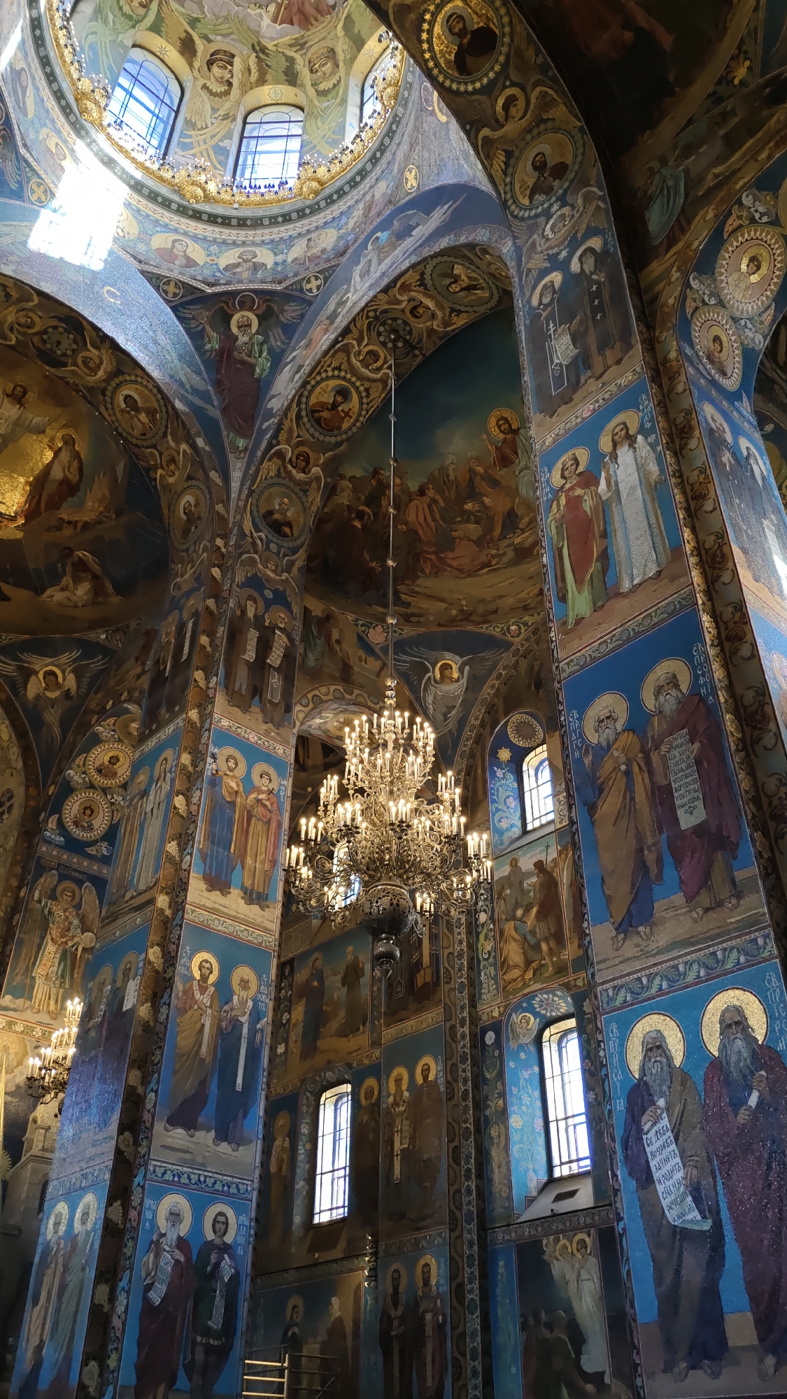 This image captures the opulent interior of a cathedral, likely in Saint Petersburg, Russia, given the location context. The scene is dominated by vibrant, detailed religious iconography covering the walls and ceilings. Numerous figures, depicted in religious robes and halos, are rendered in mosaics and frescoes. These figures include saints, angels, and possibly biblical narratives.
The central focus of the image is a large, ornate chandelier, its numerous lit candles casting a warm glow throughout the space. Sunlight streams in from arched windows, illuminating parts of the dome and contributing to the ethereal atmosphere. The architectural elements, such as the vaulted ceilings and soaring columns, are richly decorated with gold accents and intricate patterns, creating a sense of grandeur and reverence.
The overall impression is one of solemnity and spiritual grandeur. The detailed artwork suggests a place of significant religious and historical importance. While no specific event is depicted, the lighting suggests it could be daytime, with natural light contributing to the overall ambiance. The pristine condition of the artworks and the functioning chandelier point to an active and well-maintained place of worship.
Visible text, though in Cyrillic script, is present on scrolls held by some of the figures, further emphasizing the religious theme. The sheer volume and intricacy of the artwork suggest a significant undertaking in its creation, indicative of a renowned religious site.