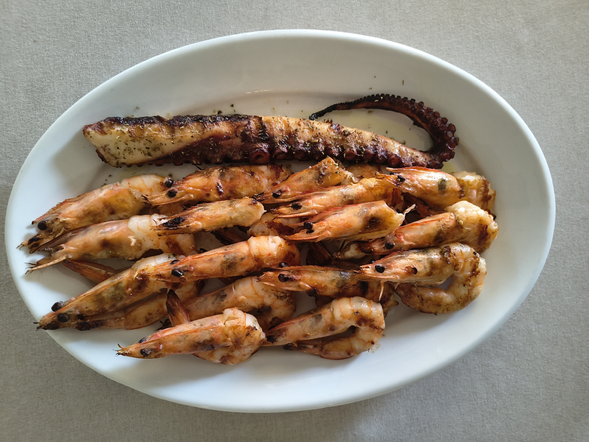 A white oval platter filled with grilled seafood sits on a light gray tablecloth. The platter contains a pile of grilled shrimp and a piece of grilled octopus tentacle. The shrimp are pinkish-orange with grill marks and some black spots. The octopus tentacle is dark brown and appears to be seasoned with herbs. The background is a plain tablecloth. The scene is captured from a top-down perspective. This image was taken in Limenaria, Greece.