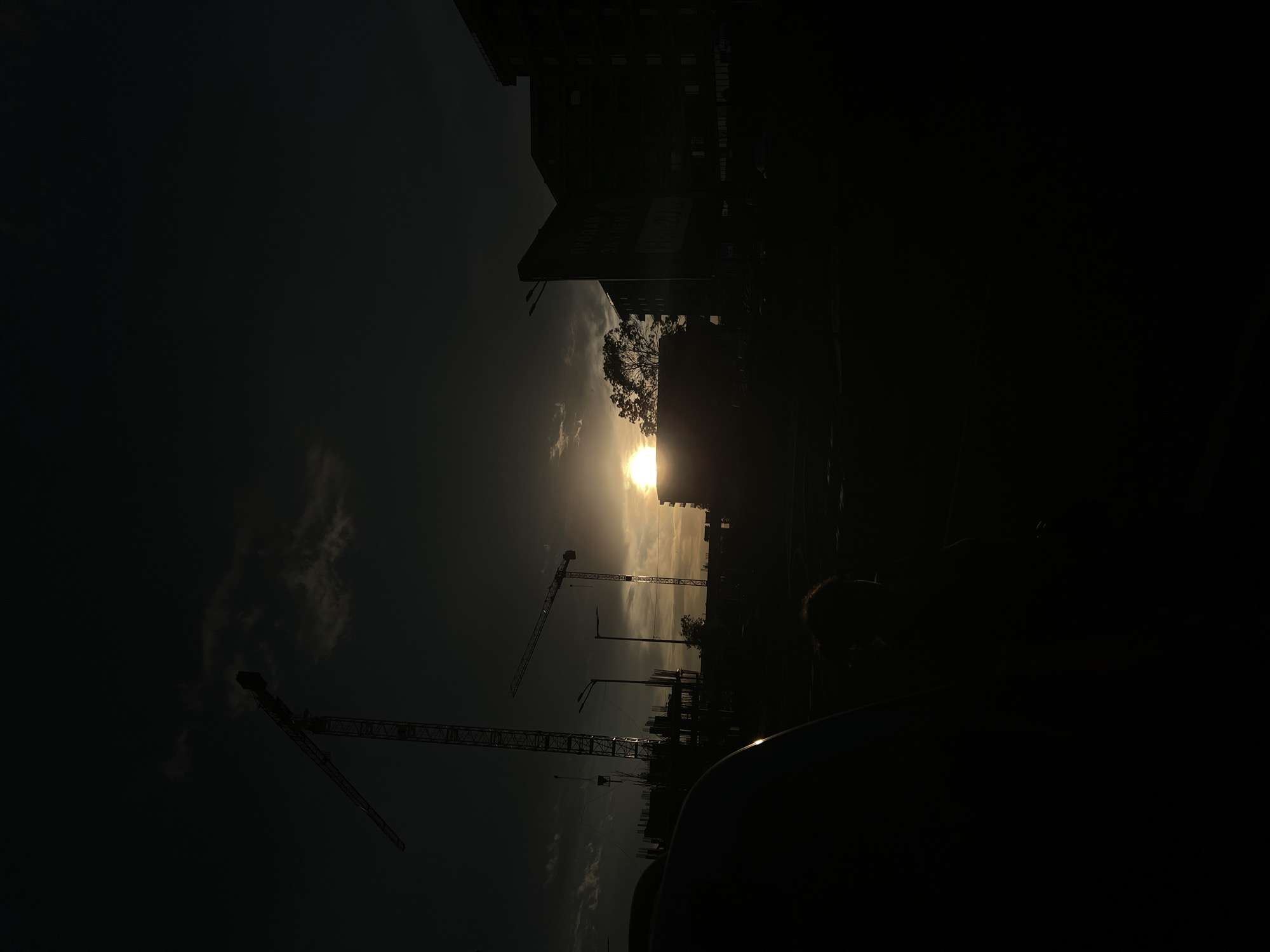 The image shows a low-angle, slightly blurred view of a construction site in Burgas, Bulgaria, at sunset. Three construction cranes are silhouetted against the bright sun, which is partially obscured by buildings and a tree. The surrounding environment includes buildings, streetlights, and a dark object in the foreground, potentially part of a vehicle's interior. The sky is dark with some visible clouds. The overall scene is dark due to the sunset and contrast.