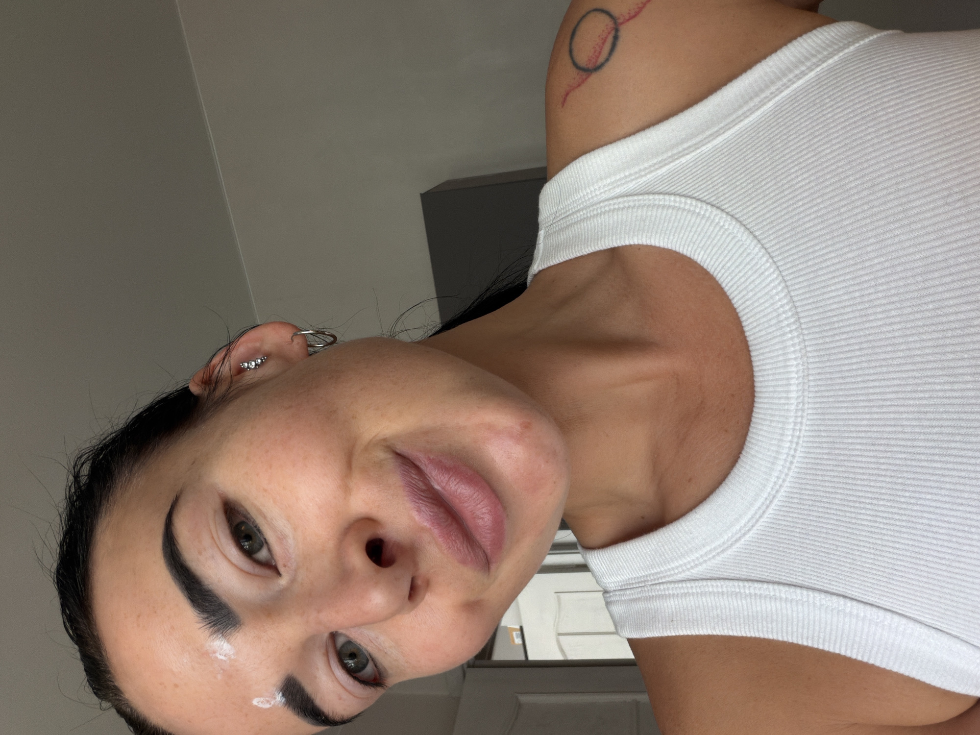 A close-up shot of a woman wearing a white tank top. She has dark hair, defined eyebrows, and multiple piercings in her left ear. A small tattoo is visible on her right shoulder. White cream is visible in the upper left of the eyebrow. The background shows a wall. There is no indication of the location being in Saint Petersburg, Russia.