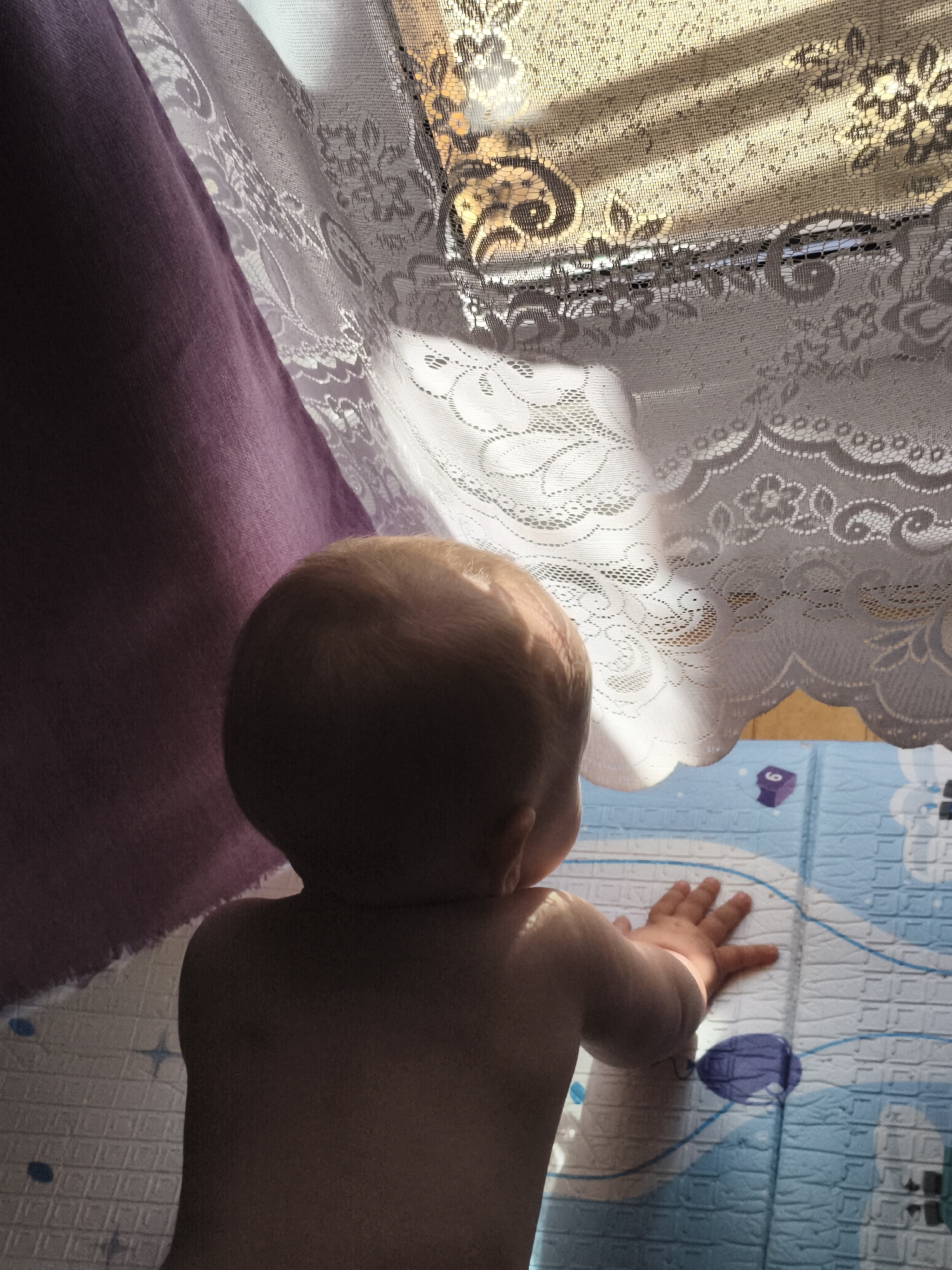 A baby, viewed from behind, rests their hands on a blue and white patterned mat. The baby's back and head are illuminated by sunlight filtering through a white lace curtain hanging on a window. To the left, a purple drape partially obscures the scene. The mat on the floor features cartoonish drawings and geometric patterns. This image was captured in Chaikovsky, Russia.