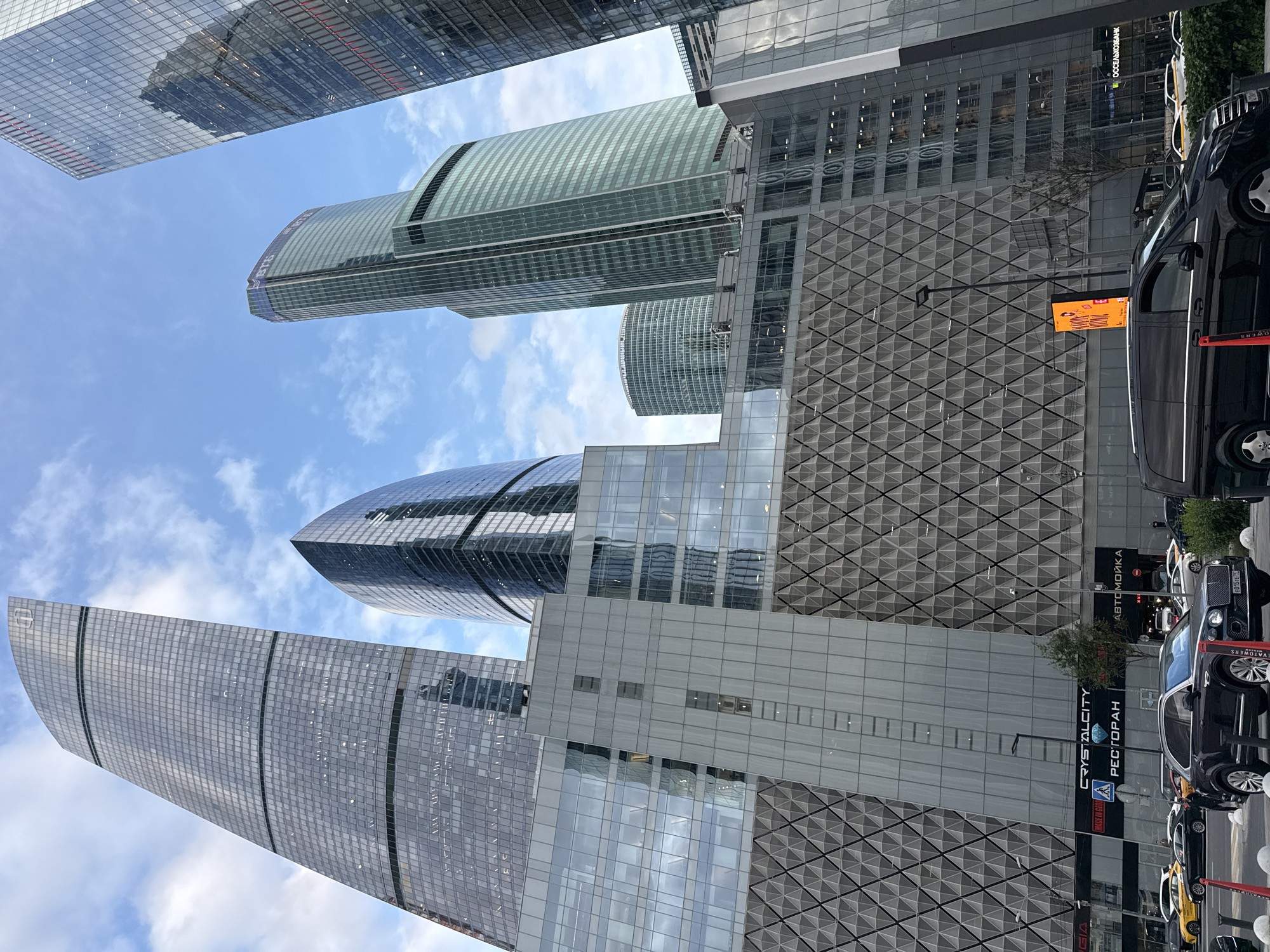 This media file captures a perspective looking upwards at a cluster of modern skyscrapers in an urban environment. The buildings are characterized by their sleek, glass facades, reflecting the sky and clouds. In the foreground, the top portion of a dark-colored van is visible, with its wheels and side door clearly depicted. Further down the street, a line of vehicles, including a black SUV, a yellow car, and other vehicles, are parked.
One of the buildings features a distinct diamond-patterned facade. Visible text on the buildings includes "CRYSTAL CITY" and "PECTOPAH" (restaurant in Russian), alongside a road sign with Russian text that appears to read "АВТОМОЙКА" (car wash). The sky is partly cloudy, suggesting it's daytime, possibly late afternoon given the lighting. The overall scene depicts a contemporary urban setting with a focus on impressive architecture and the presence of vehicles. There are no discernible people in the image. The angle suggests the photograph was taken from street level, looking up. The location is identified as Khimki, Russia.