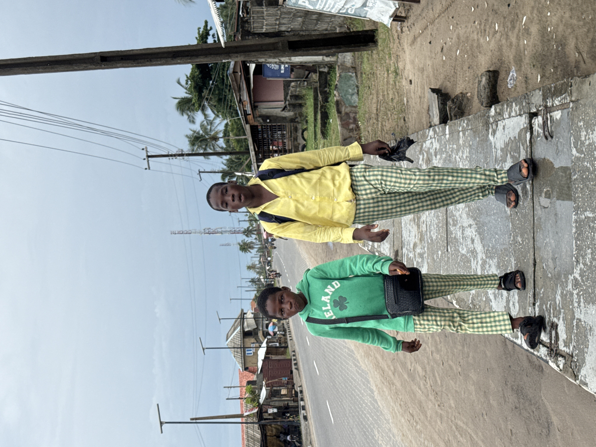 Two young men stand on a sidewalk near a road. The man on the right wears a yellow shirt and plaid pants with sandals. The man on the left wears a green sweatshirt with "Ireland" on it, plaid pants, and sandals. The road behind them has a few vehicles on it and is lined with utility poles and structures that appear to be shops or homes. The background shows palm trees and a cloudy sky. The scene appears to be in a city or town setting, possibly in Badagry, Nigeria, based on the visual clues of the environment.