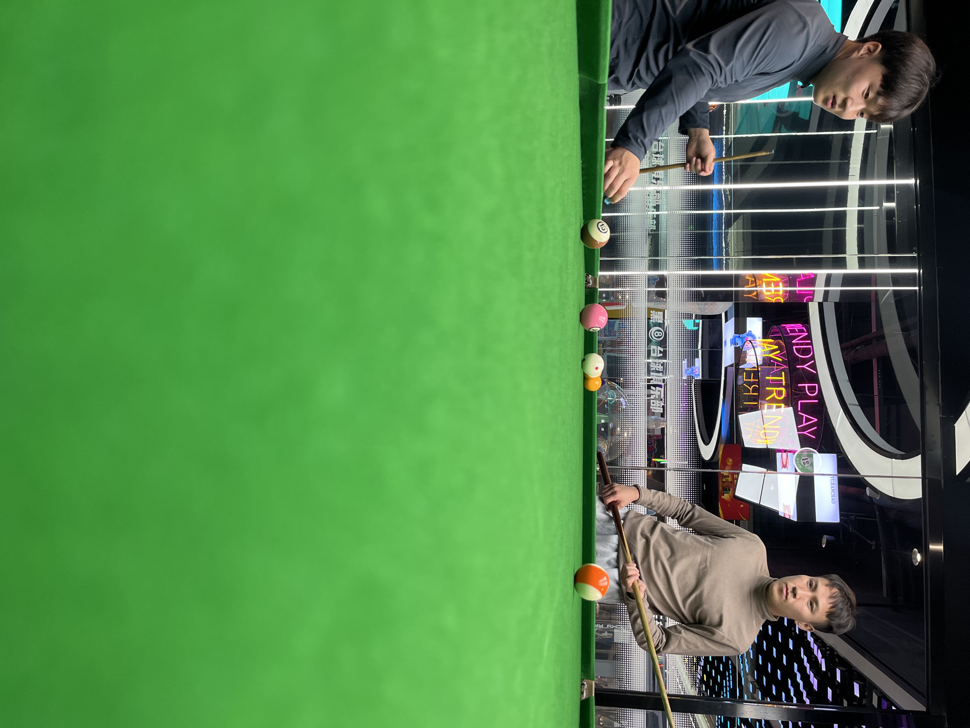 The image shows two young men playing billiards in an indoor setting. One man, wearing a grey long-sleeved shirt, is positioned on the right side of the frame, holding a pool cue and aiming at a ball on the green felt table. Several other billiard balls are visible on the table, including a striped ball and solid colored balls. The other man, wearing a beige turtleneck and grey pants, is on the lower right, also holding a pool cue. He appears to be in the process of striking a ball. The background features a brightly lit environment with multiple neon signs displaying text in English and Chinese, suggesting a recreational or entertainment venue. Some of the visible text includes "TRENDY PLAY," "YIJIAOHAO," and Chinese characters that translate to "Black and White Entertainment." The overall ambiance is lively and modern, with reflections in the glass surfaces adding to the visual complexity of the scene. The lighting suggests it is likely evening or indoors with artificial lighting. The men appear focused on the game. The location is identified as Daxing District, China.