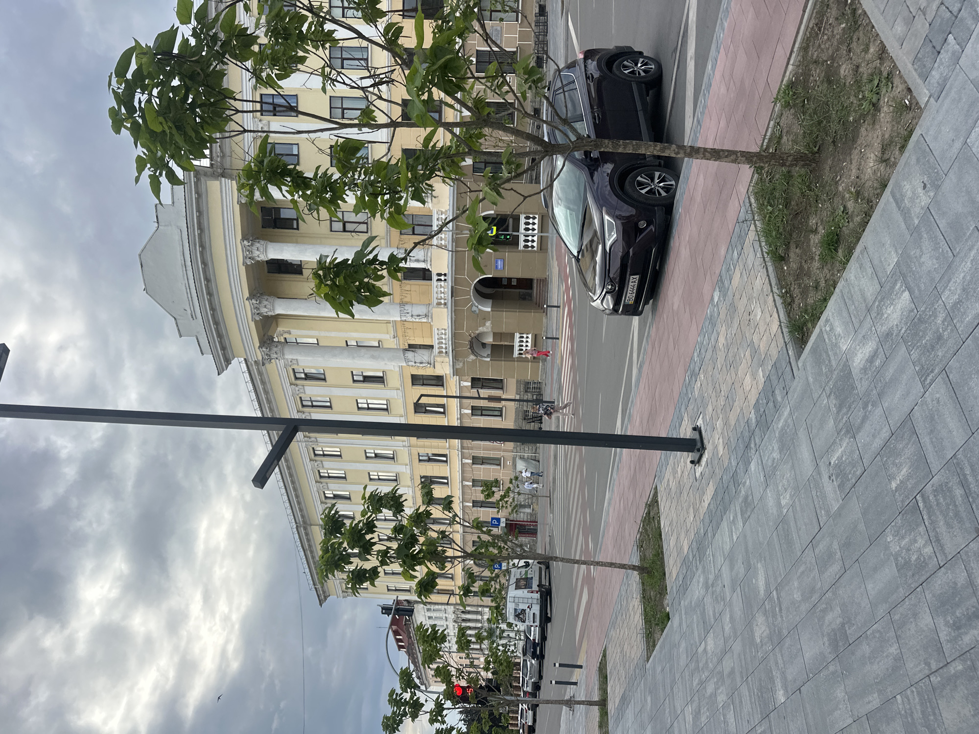 The image depicts a street scene in Ternopil, Ukraine, likely in front of a grand, light-yellow building with classical architectural elements such as columns and an arched entryway. The sky is overcast with gray clouds. In the foreground, a pedestrian walkway paved with gray and pinkish-gray tiles leads to a street. Several young trees with green foliage are planted along the sidewalk. A dark-colored SUV is parked on the right side of the street, partially obscuring a tree. Further down the street, there are other cars, a white van, and a traffic light showing red. Pedestrians are visible on the sidewalk and crossing the street at a marked crosswalk. The building's facade features multiple windows and decorative moldings. The overall impression is a well-maintained urban environment on a cloudy day.