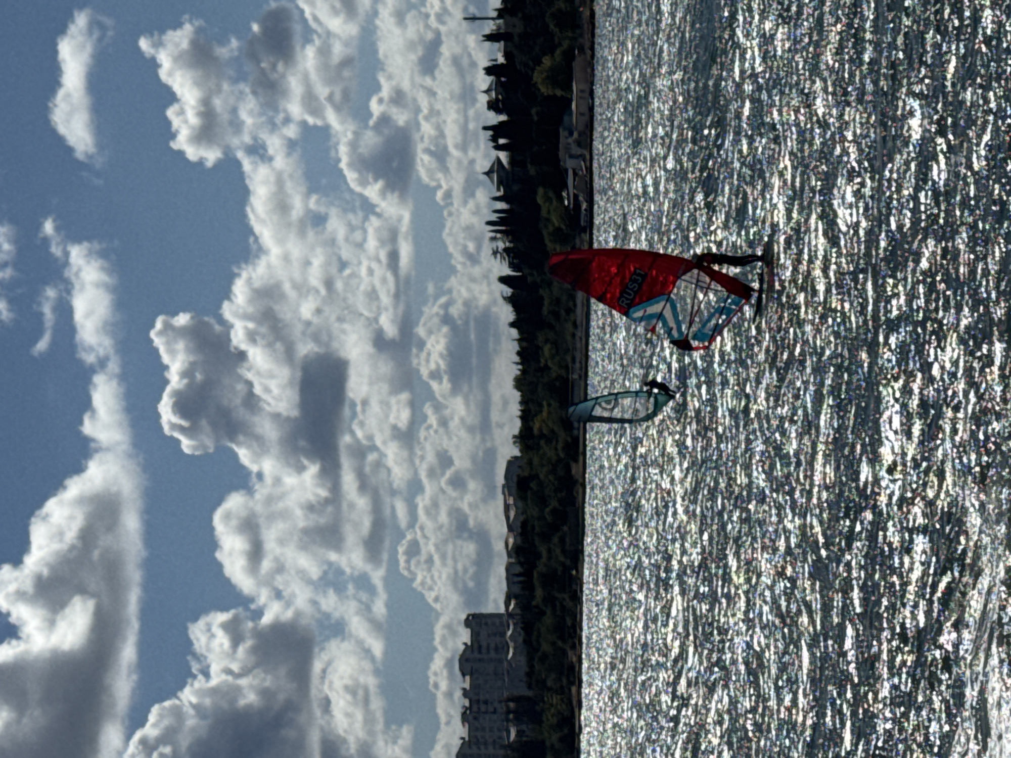 Two windsurfers are on the water with a cityscape in the background. The water is choppy with sunlight glinting off the surface. The foreground windsurfer has a red, white, and blue sail with "RUS31" printed on it. The second windsurfer, further in the background, has a teal sail. In the distance, there is a line of trees with buildings and domes visible above them. The sky is blue with scattered, fluffy white clouds.