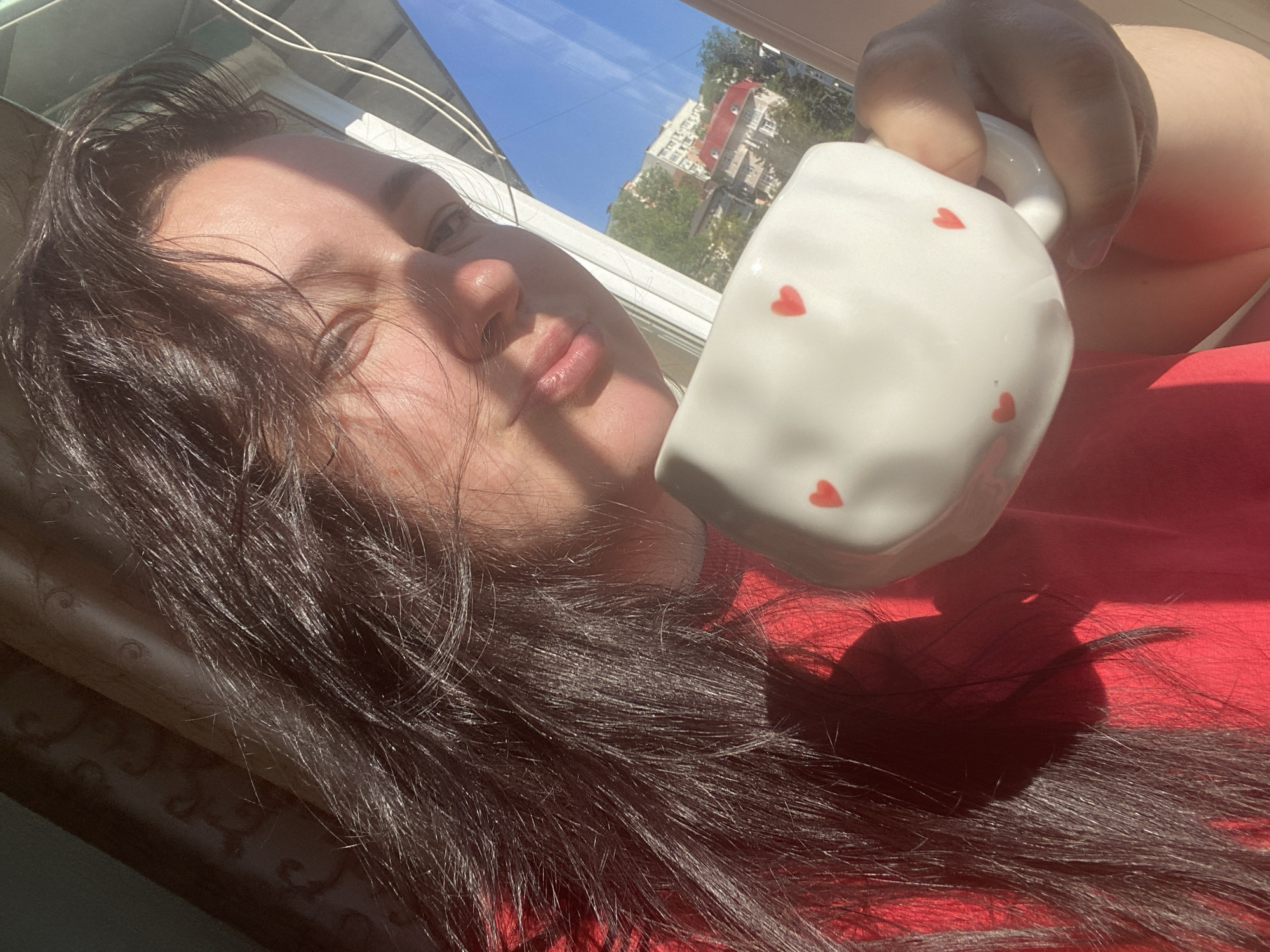 A selfie-style photograph depicts a woman holding a white ceramic mug adorned with red heart decals. The mug is positioned prominently in the foreground, obscuring parts of the woman's face and body. The woman has dark hair that cascades across a red garment, likely a shirt or blanket. Her face is partially illuminated by natural light, and her expression is neutral, with slightly pursed lips.
In the background, a window offers a view of a city landscape. The architecture suggests an urban setting, with buildings partially visible against a blue sky. Given the query's specification, this location is assumed to be Sochi, Russia. The angle of the shot indicates that the photo was taken indoors, likely near a window, during daytime hours.