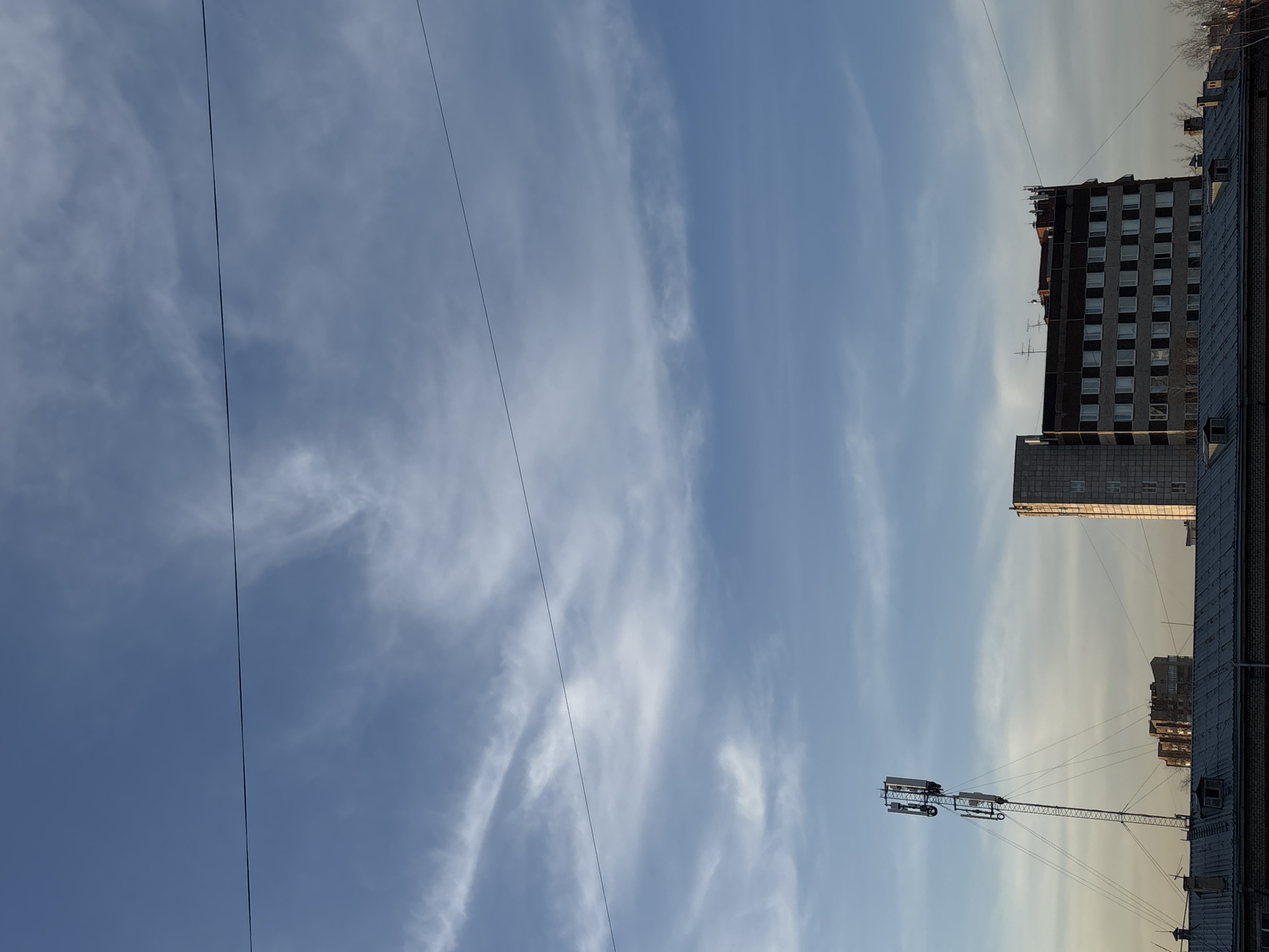 The image shows a view in Perm, Russia, oriented vertically. The scene depicts a bright blue sky with wispy clouds. Two electrical wires run diagonally across the upper left portion of the frame. Buildings are visible in the lower right and upper right portions of the image. In the lower right, a rooftop supports a cell tower. The building on the upper right is multi-story and gray. The image captures an urban environment. No people are visible.